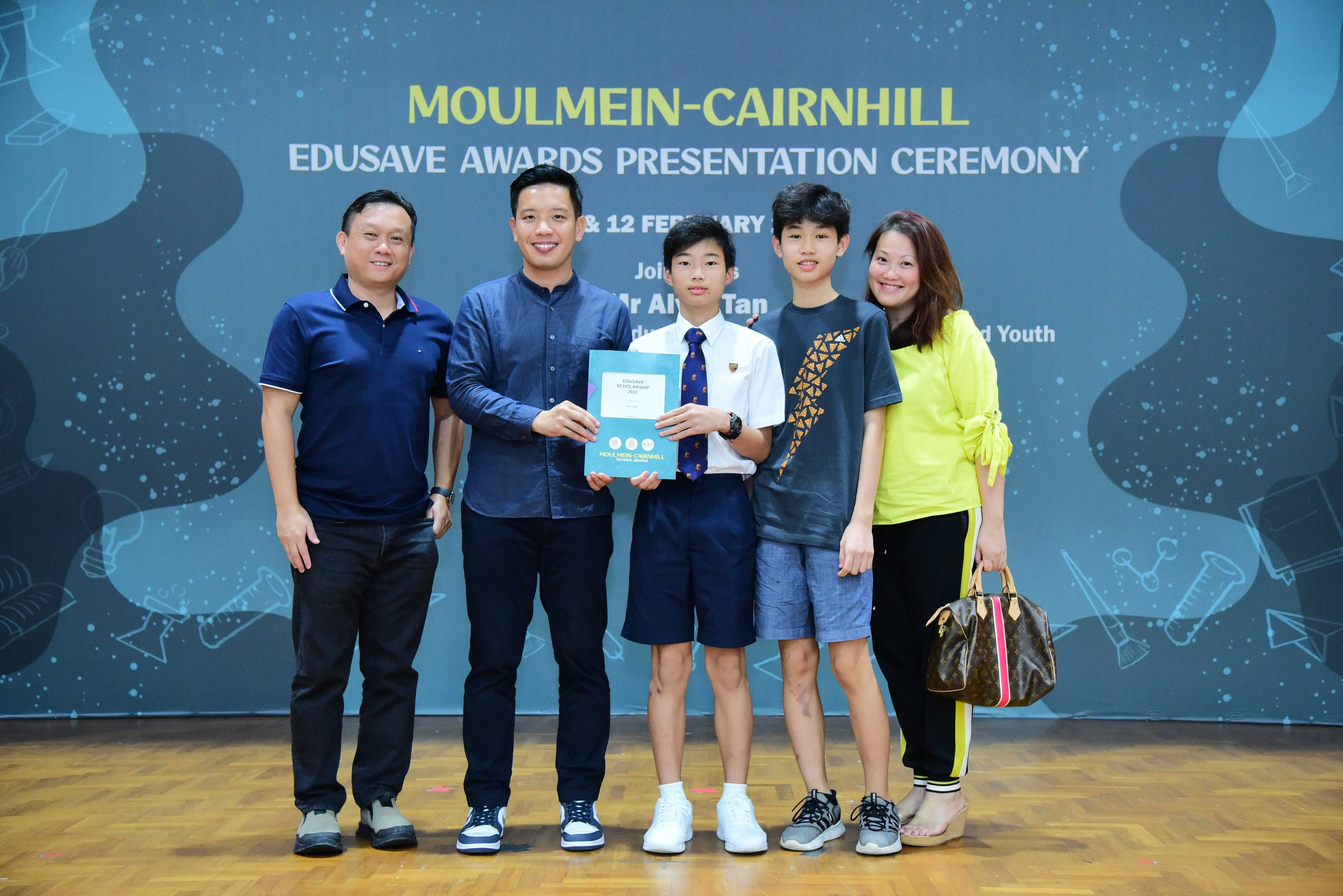 Five people pose at the Moulmein-Cairnhill Edusave Awards Presentation Ceremony, with a student holding a certificate.