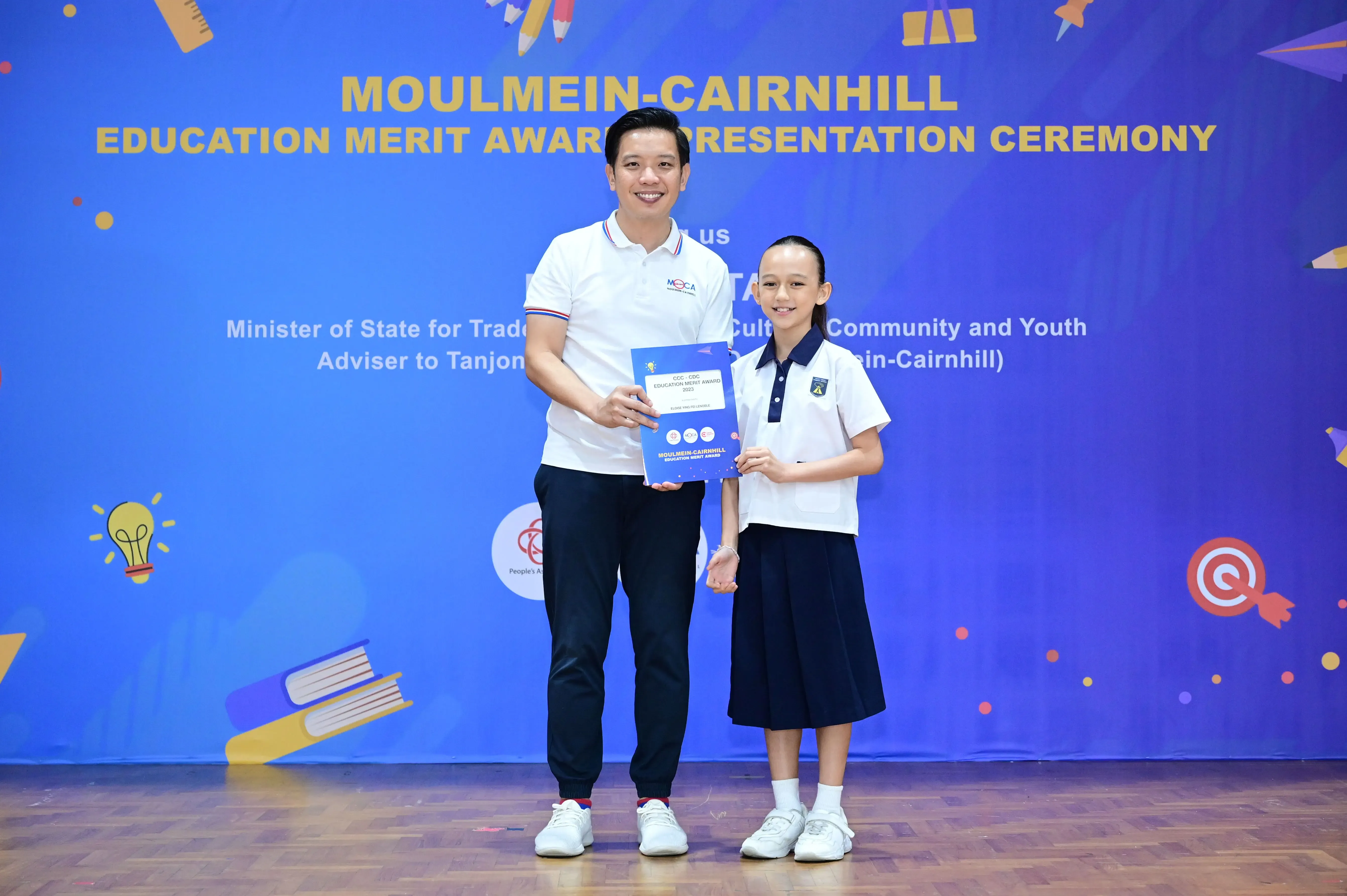 A man and a young student in uniform hold an award certificate at the Moulmein-Cairnhill Education Merit Award ceremony.