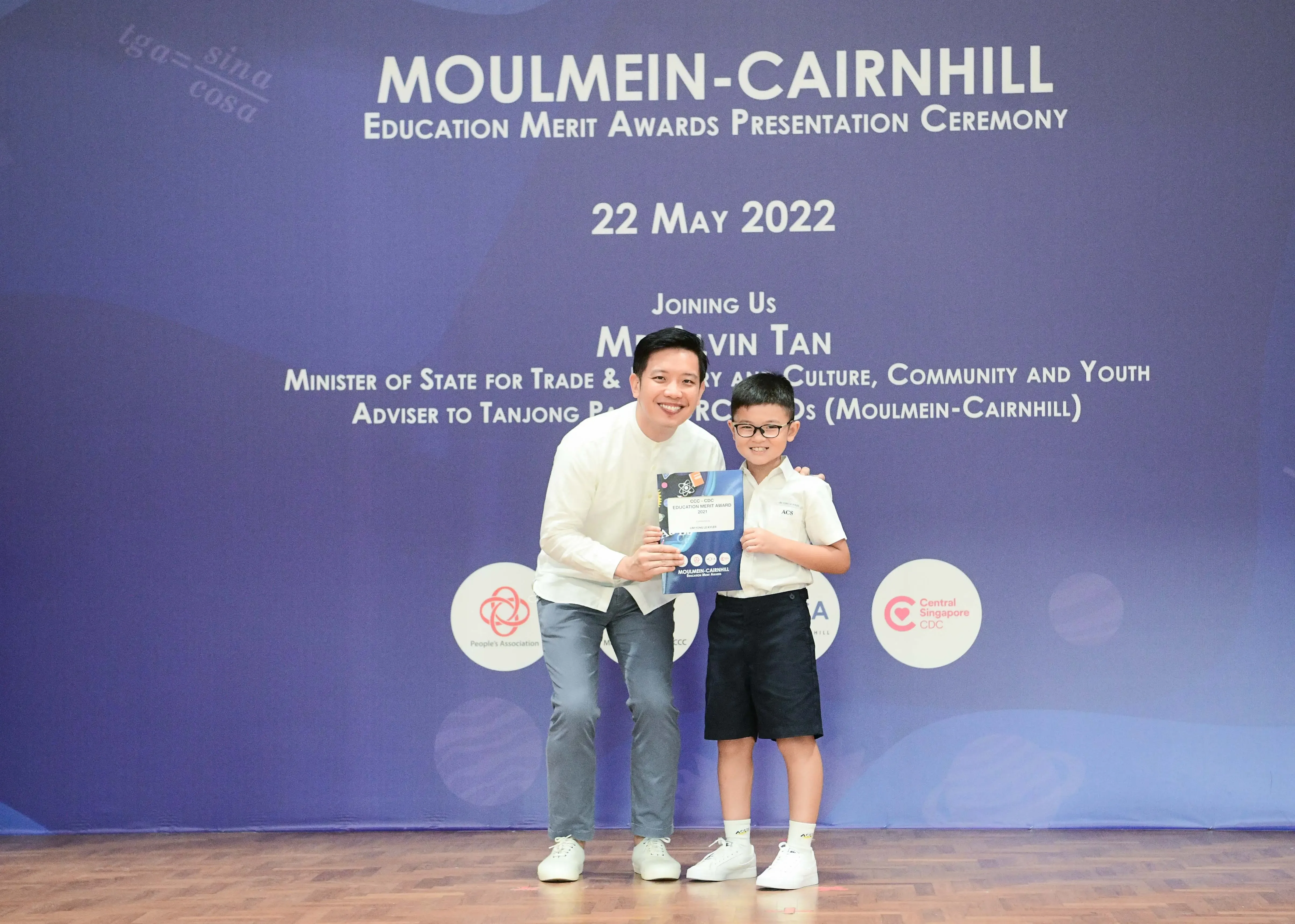 Man and young boy holding a certificate on stage at the Moulmein-Cairnhill Education Merit Awards, 22 May 2022.