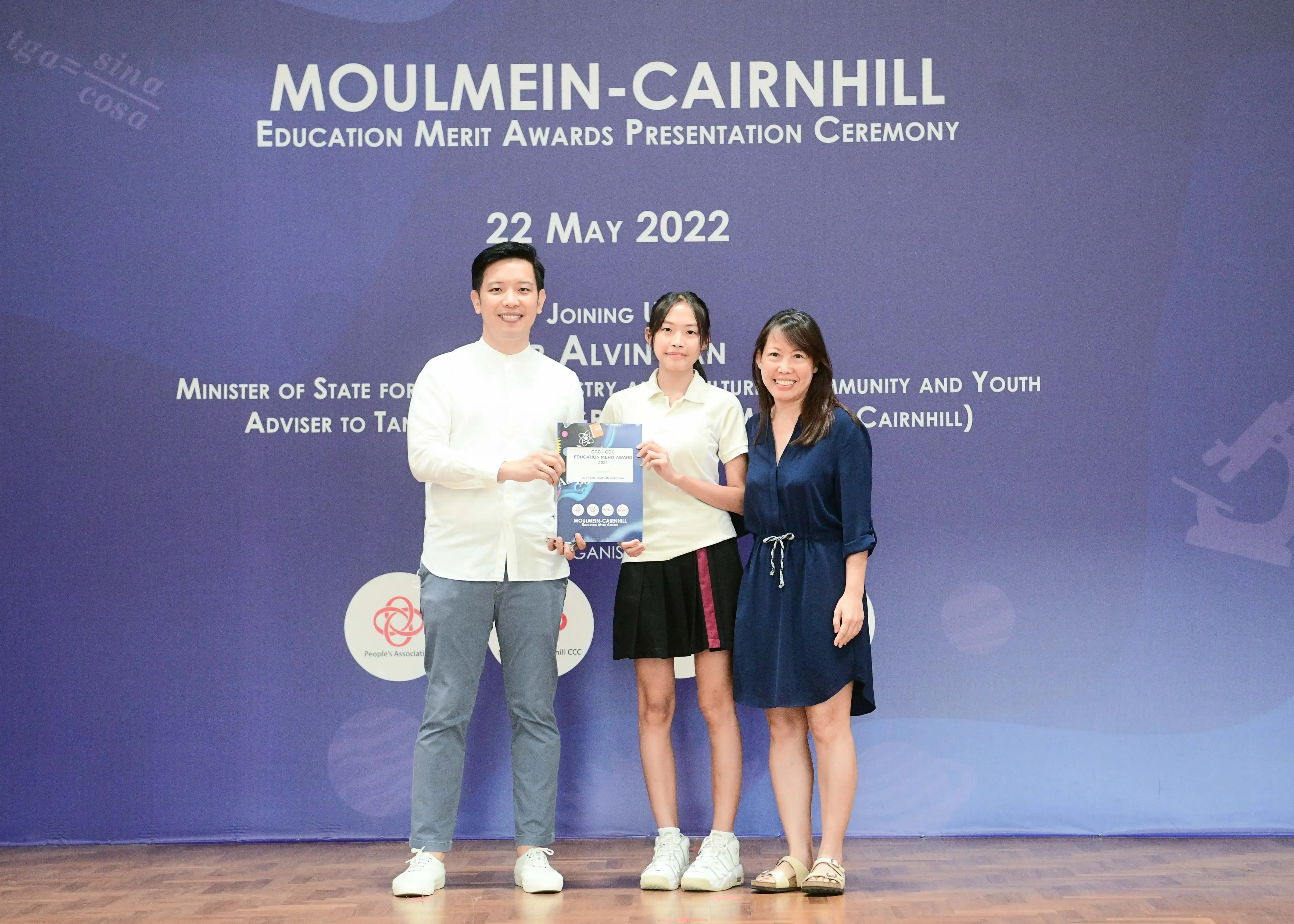 Three people stand on a stage at the Moulmein-Cairnhill Education Merit Awards Ceremony on 22 May 2022.
