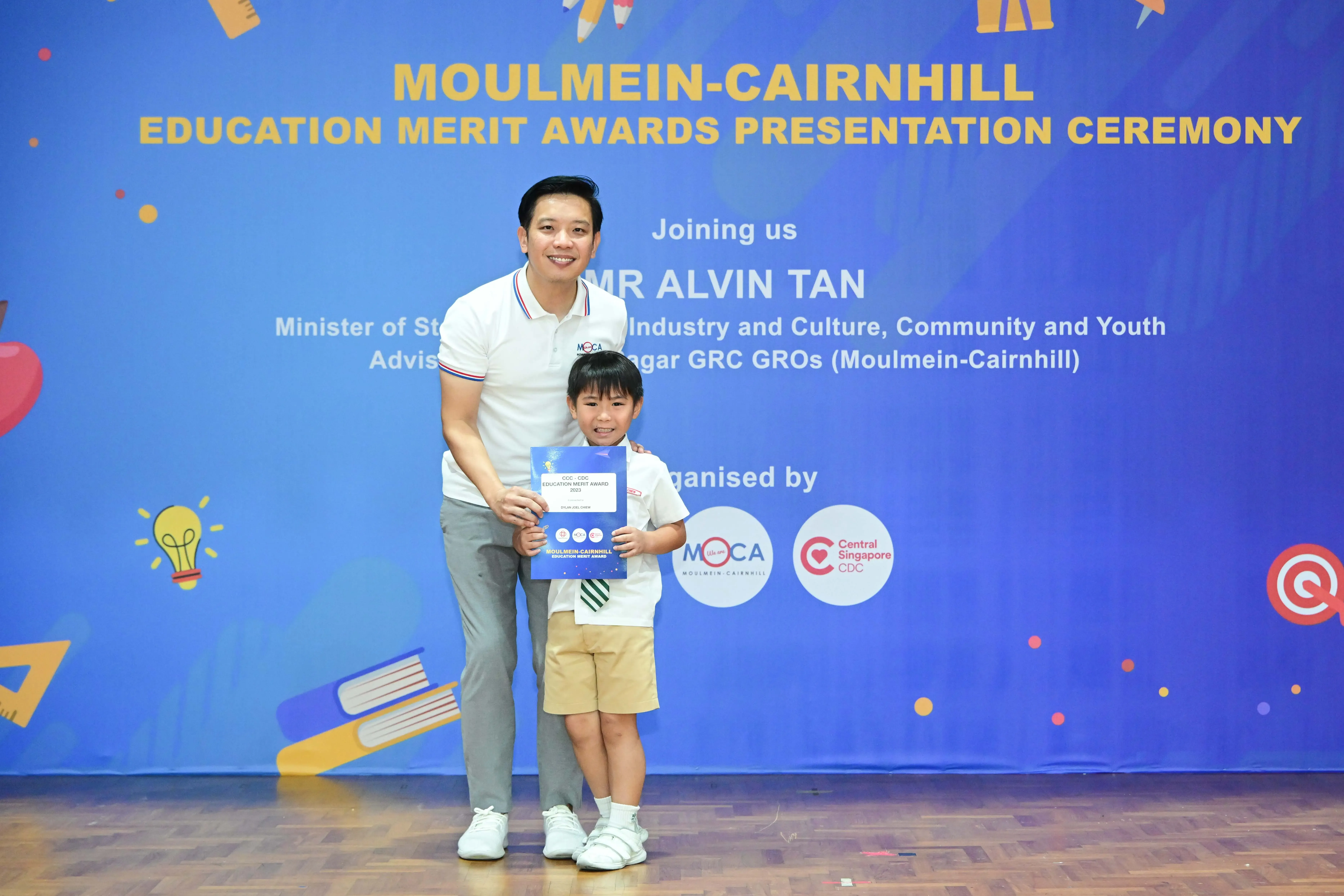 A man and young boy holding a certificate stand before a Moulmein-Cairnhill Education Merit Awards banner.
