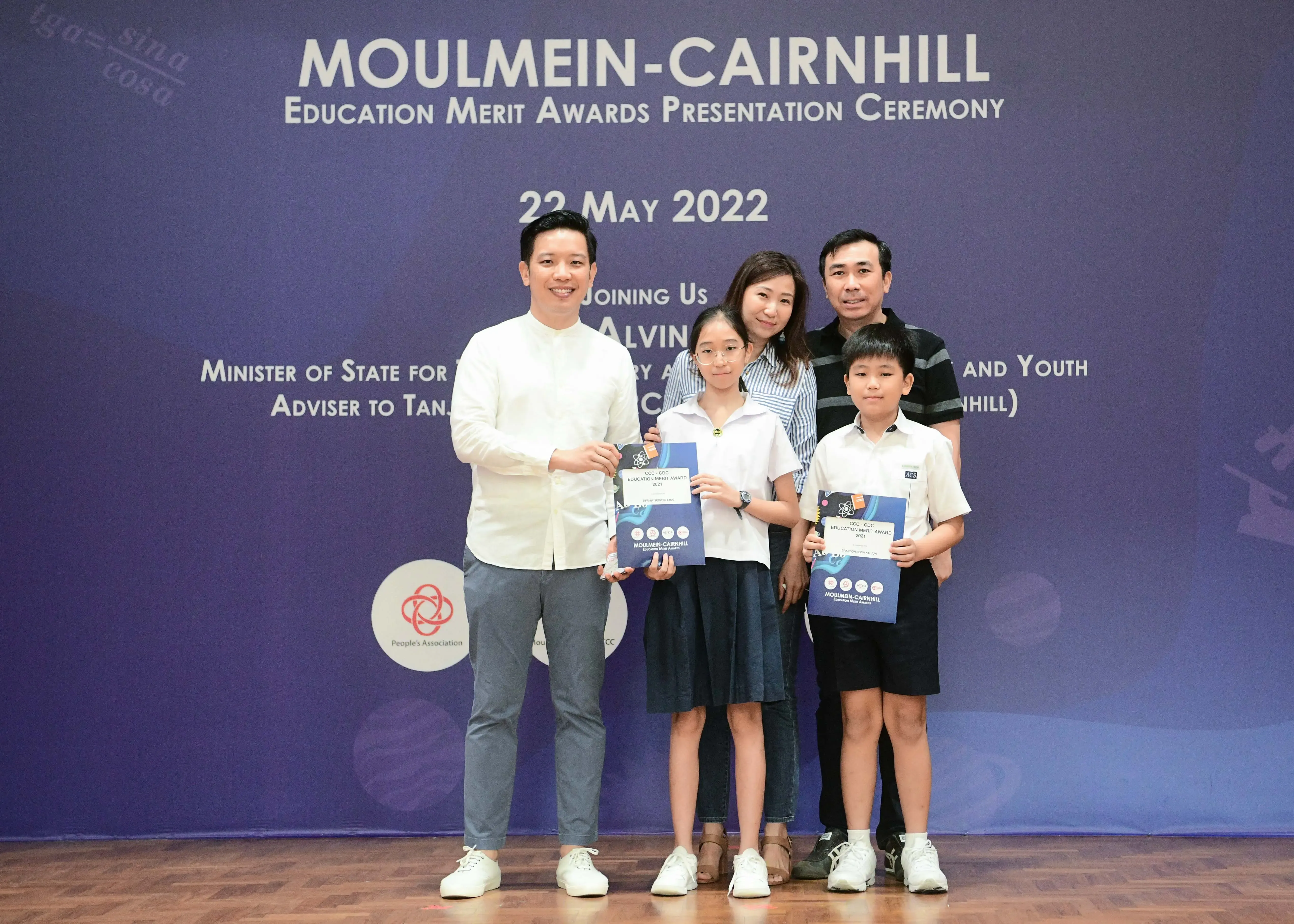 Five people pose before a Moulmein-Cairnhill Education Merit Awards banner dated 22 May 2022, two children holding award booklets.