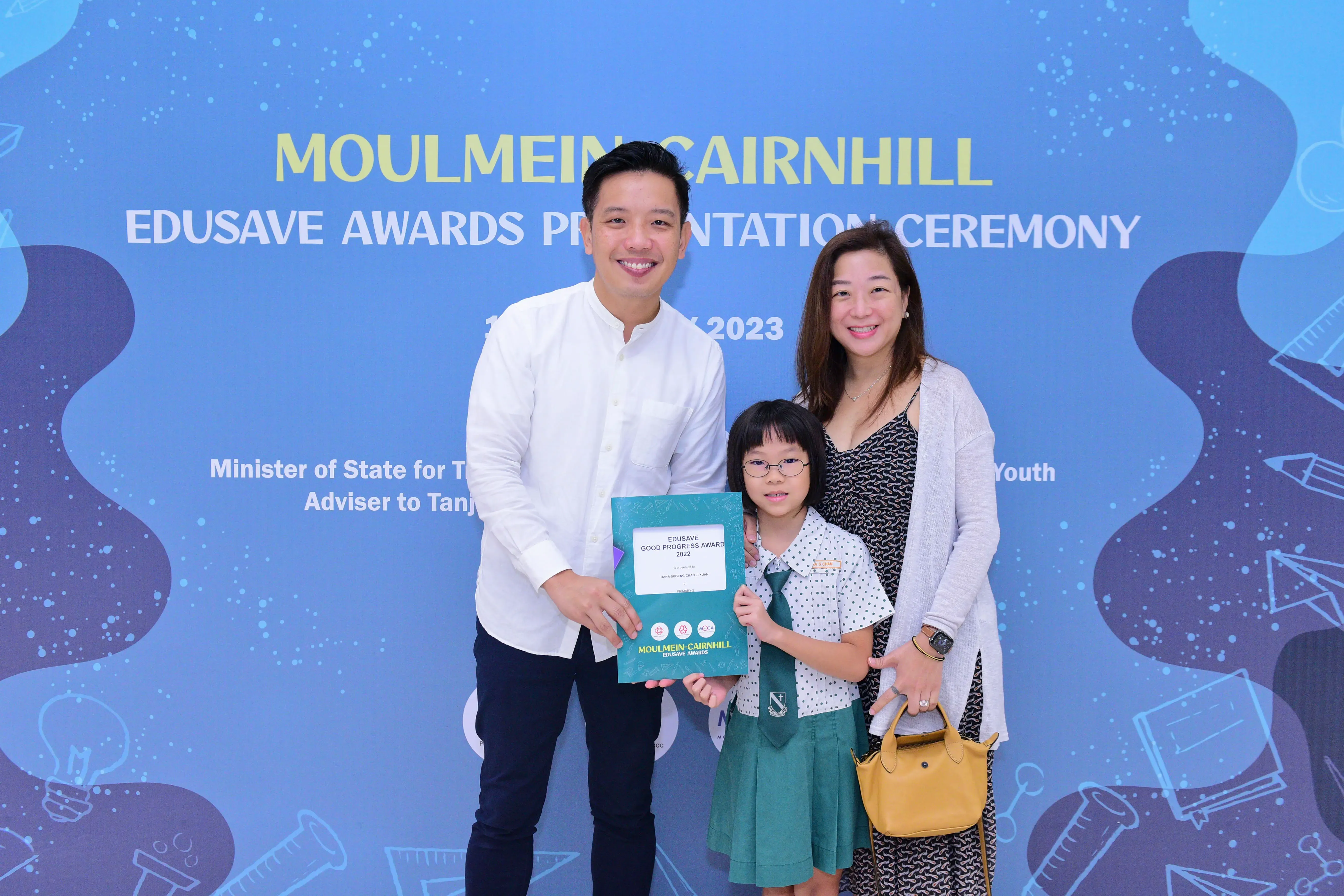 Two adults and a young girl in school uniform hold an award certificate at the Moulmein-Cairnhill Edusave Awards Ceremony 2023.
