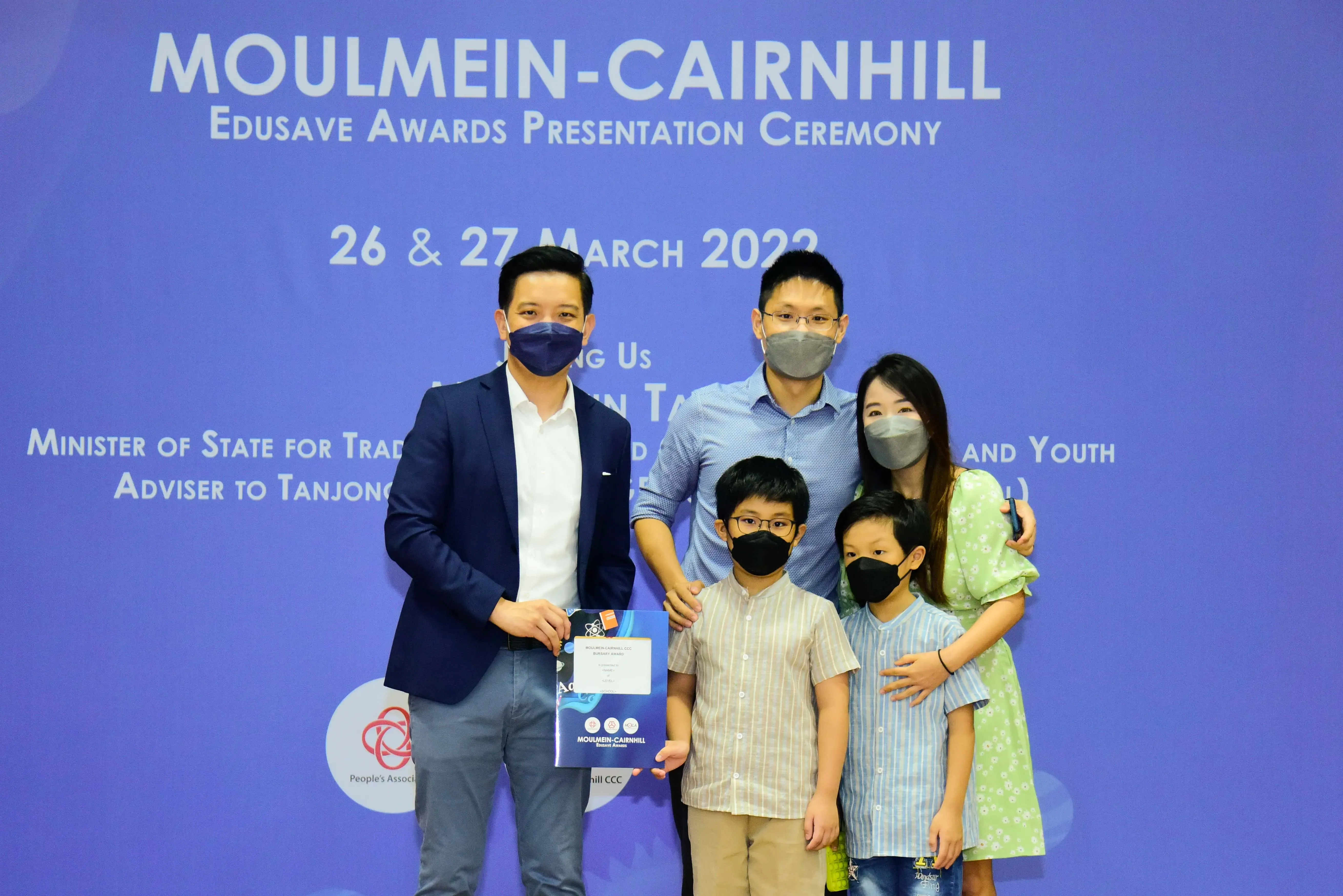 Five masked people pose before a Moulmein-Cairnhill Edusave Awards Presentation Ceremony banner dated March 2022.