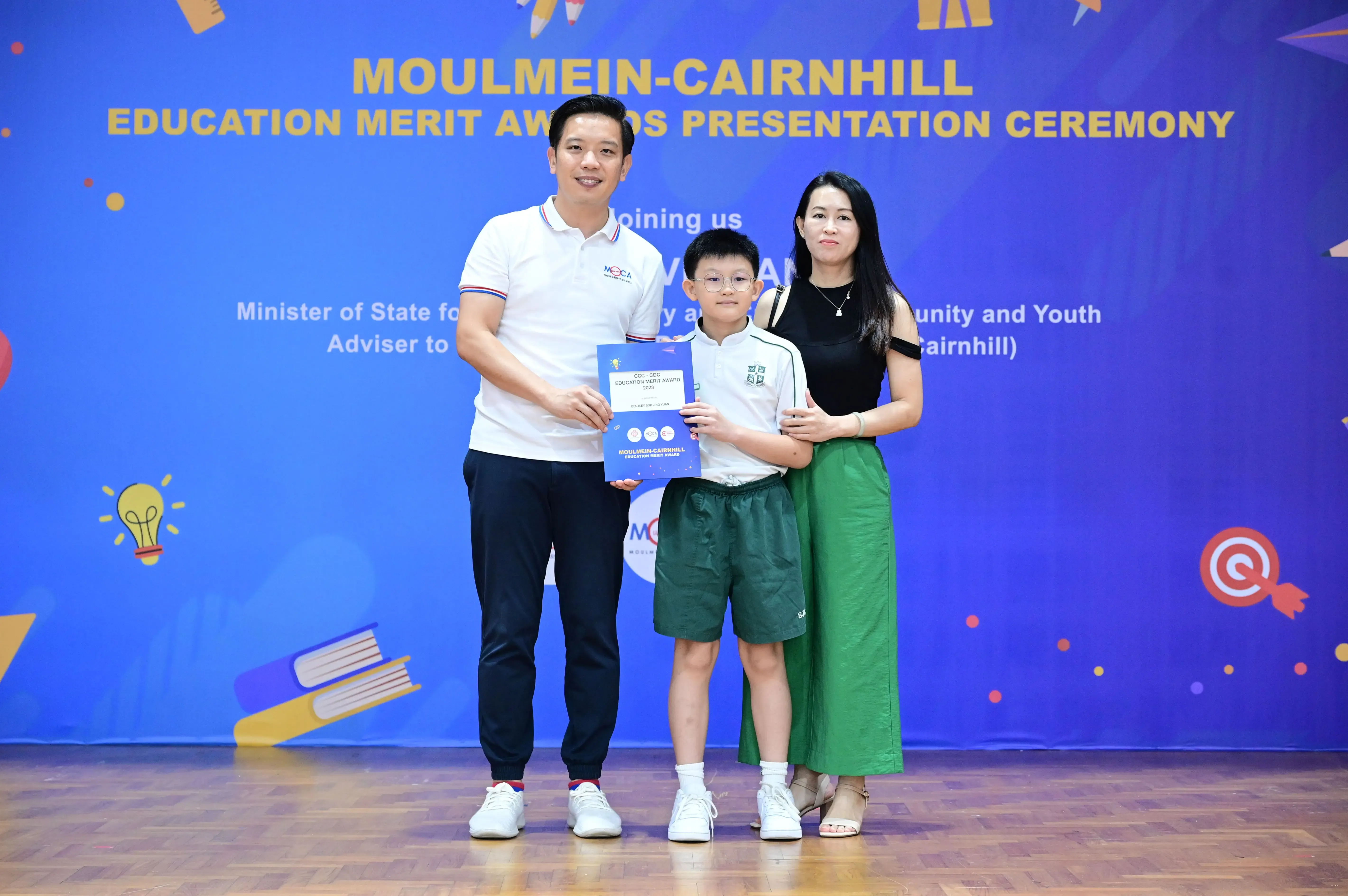 A boy holding an award certificate stands between a man and a woman at the Moulmein-Cairnhill Education Merit Awards ceremony.
