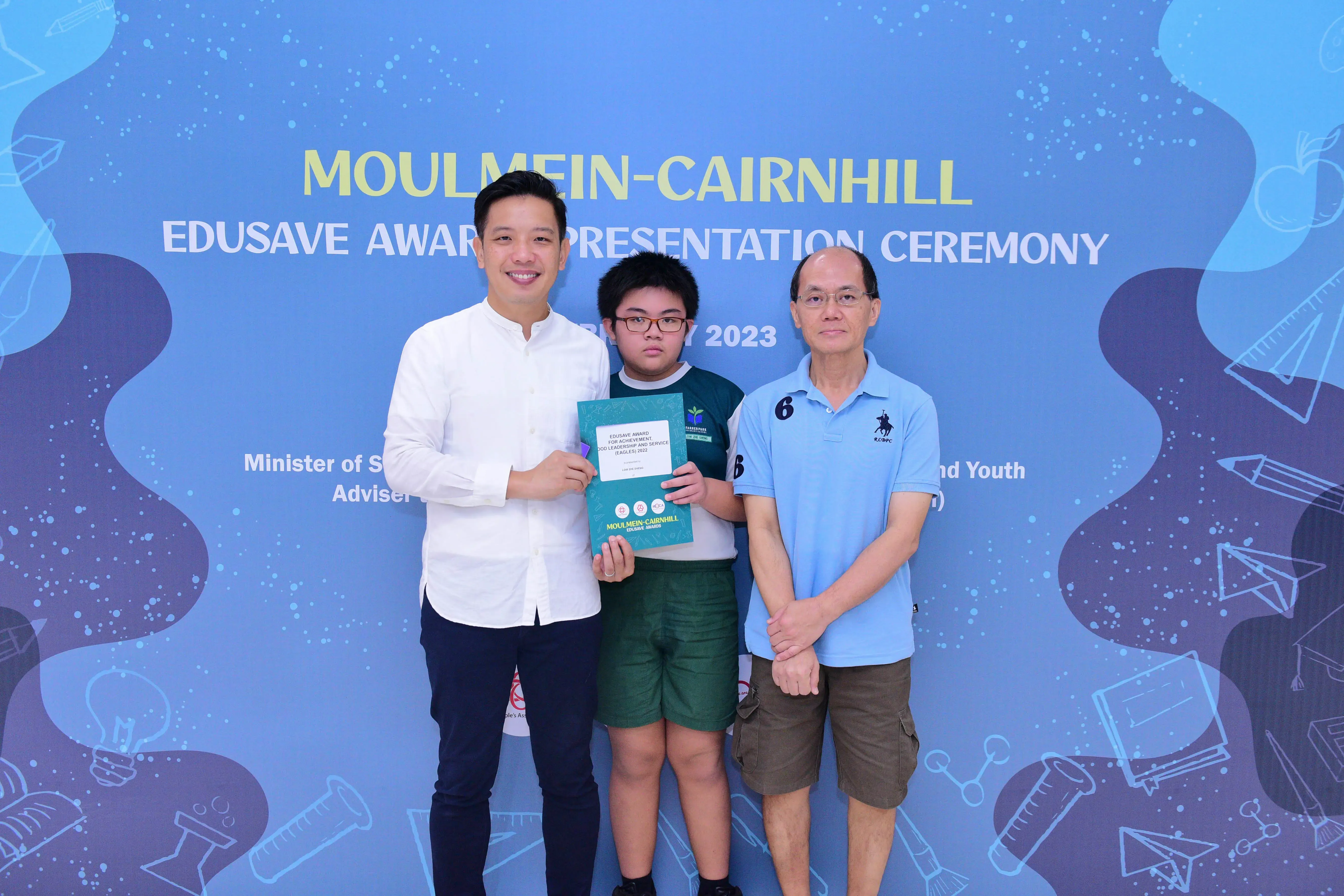 Three people pose before a Moulmein-Cairnhill Edusave Award Presentation Ceremony 2023 banner, child holds a certificate.