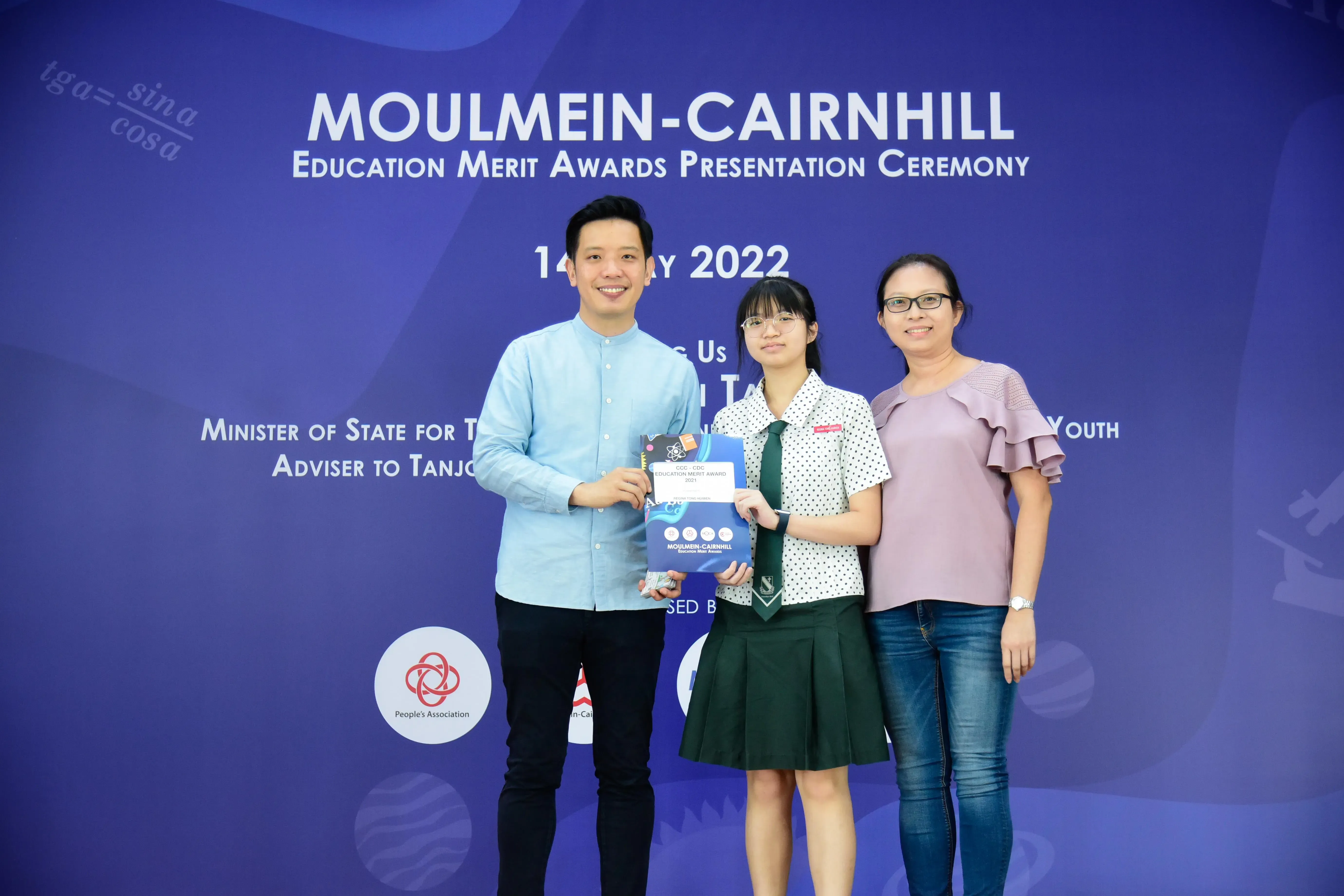 Three people smile before a Moulmein-Cairnhill Education Merit Awards 2022 banner, with a student holding an award.