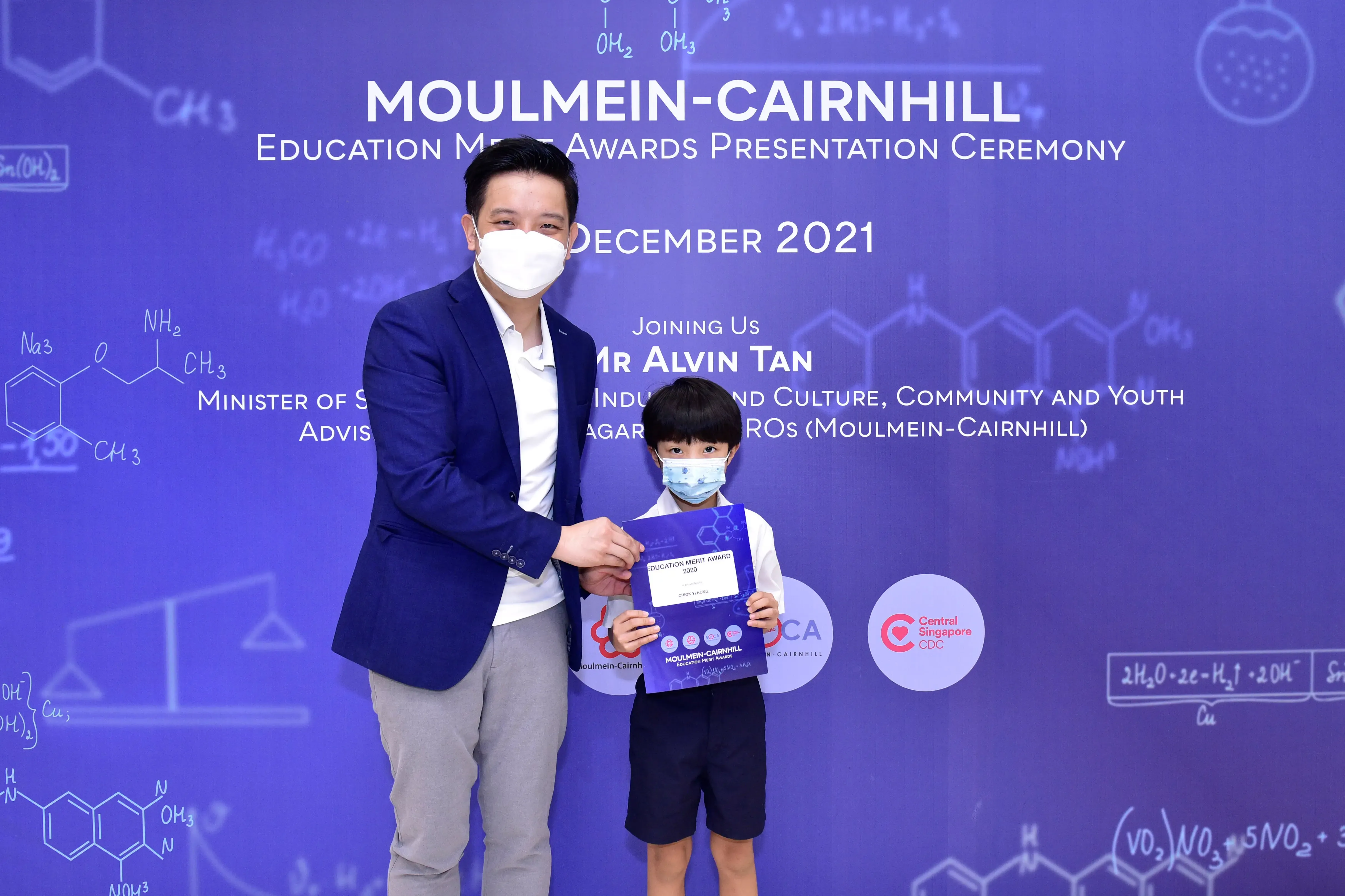 An adult and a young boy wearing masks hold an award certificate at the Moulmein-Cairnhill Education Merit Awards ceremony.