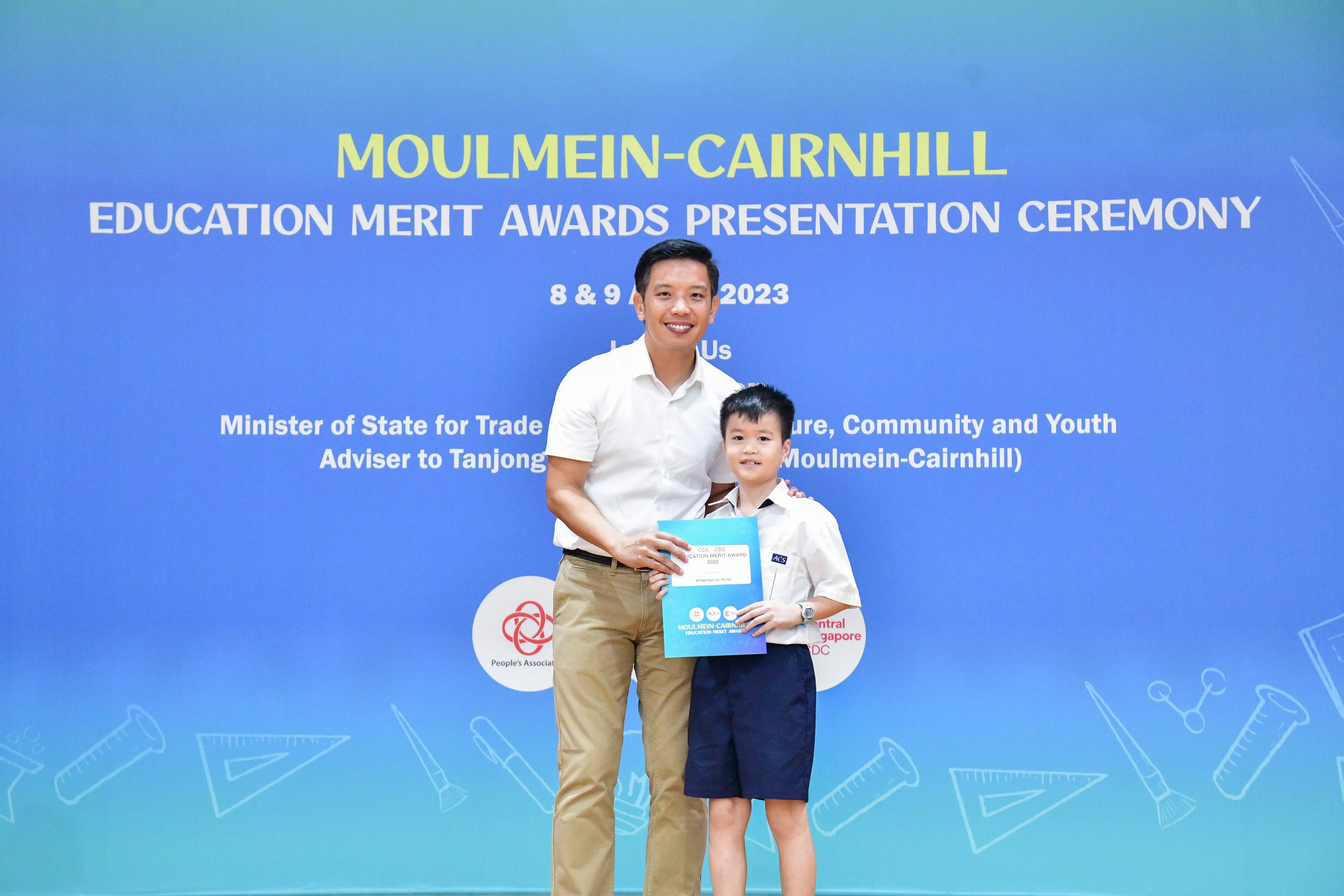 A man and a young boy in school uniform hold a certificate in front of a Moulmein-Cairnhill Education Merit Awards banner.