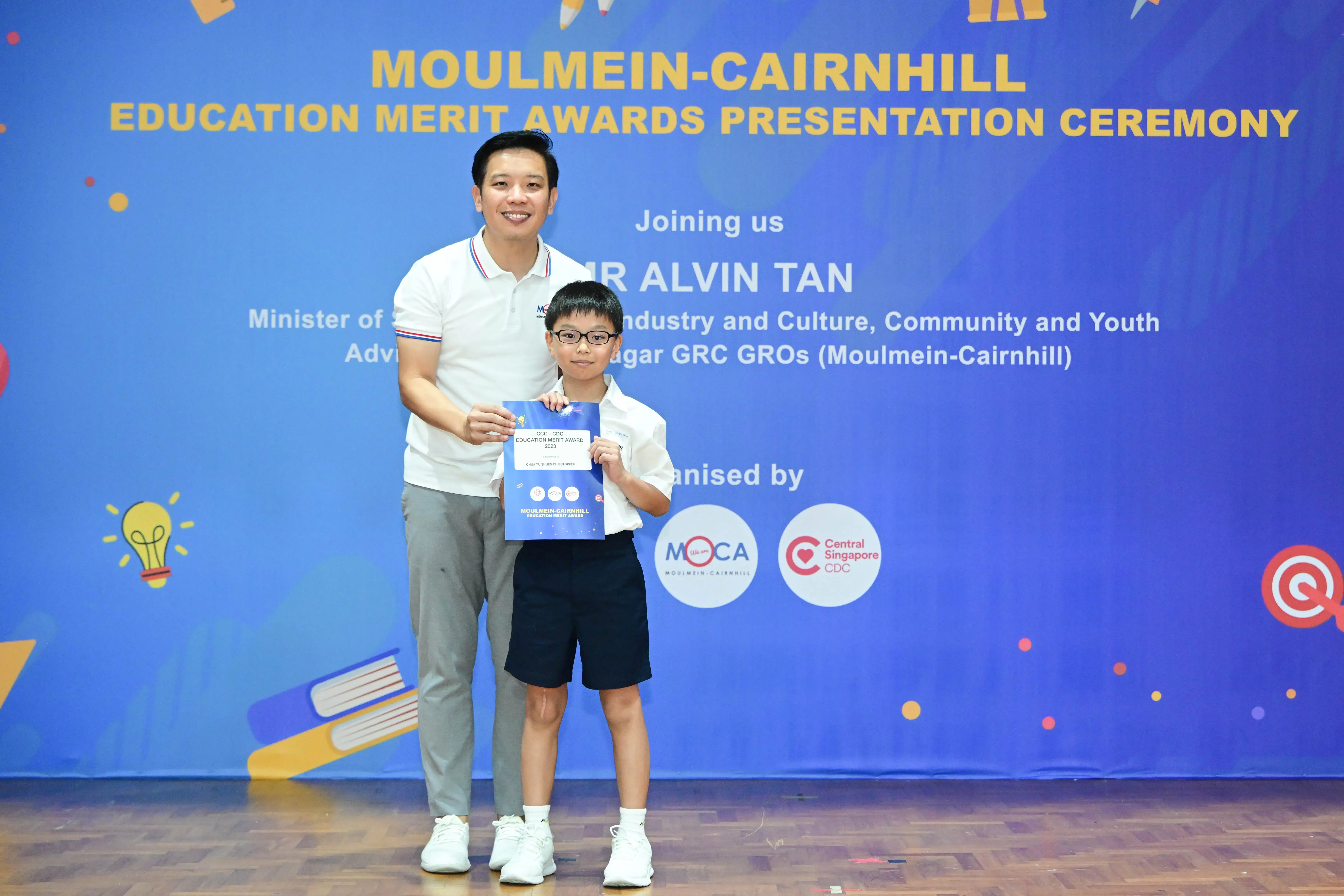 An adult and a young boy hold a blue certificate in front of a Moulmein-Cairnhill Education Merit Awards banner.