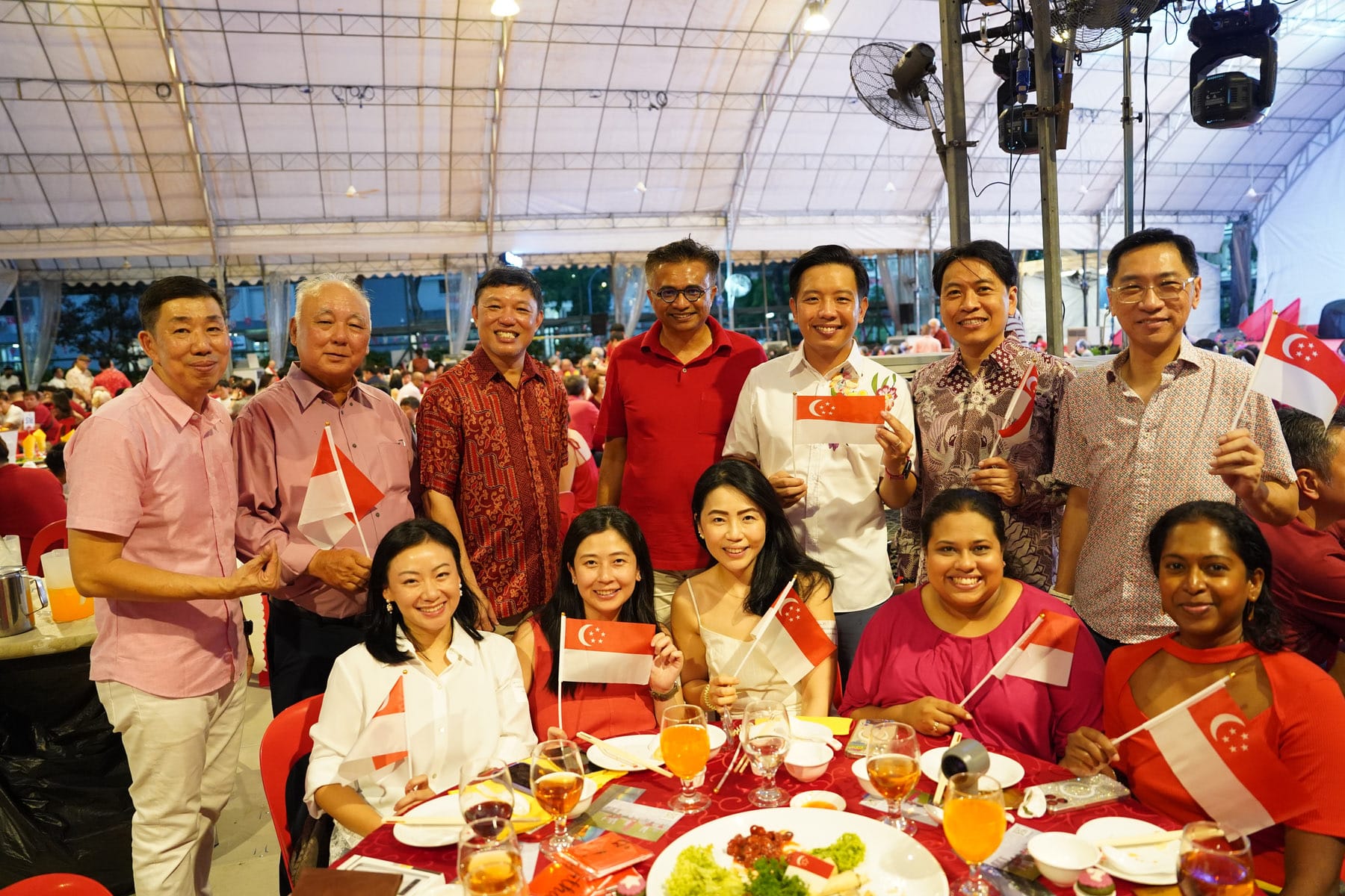 A group of smiling people holding small Singapore flags at a festive outdoor dinner under a large tent.