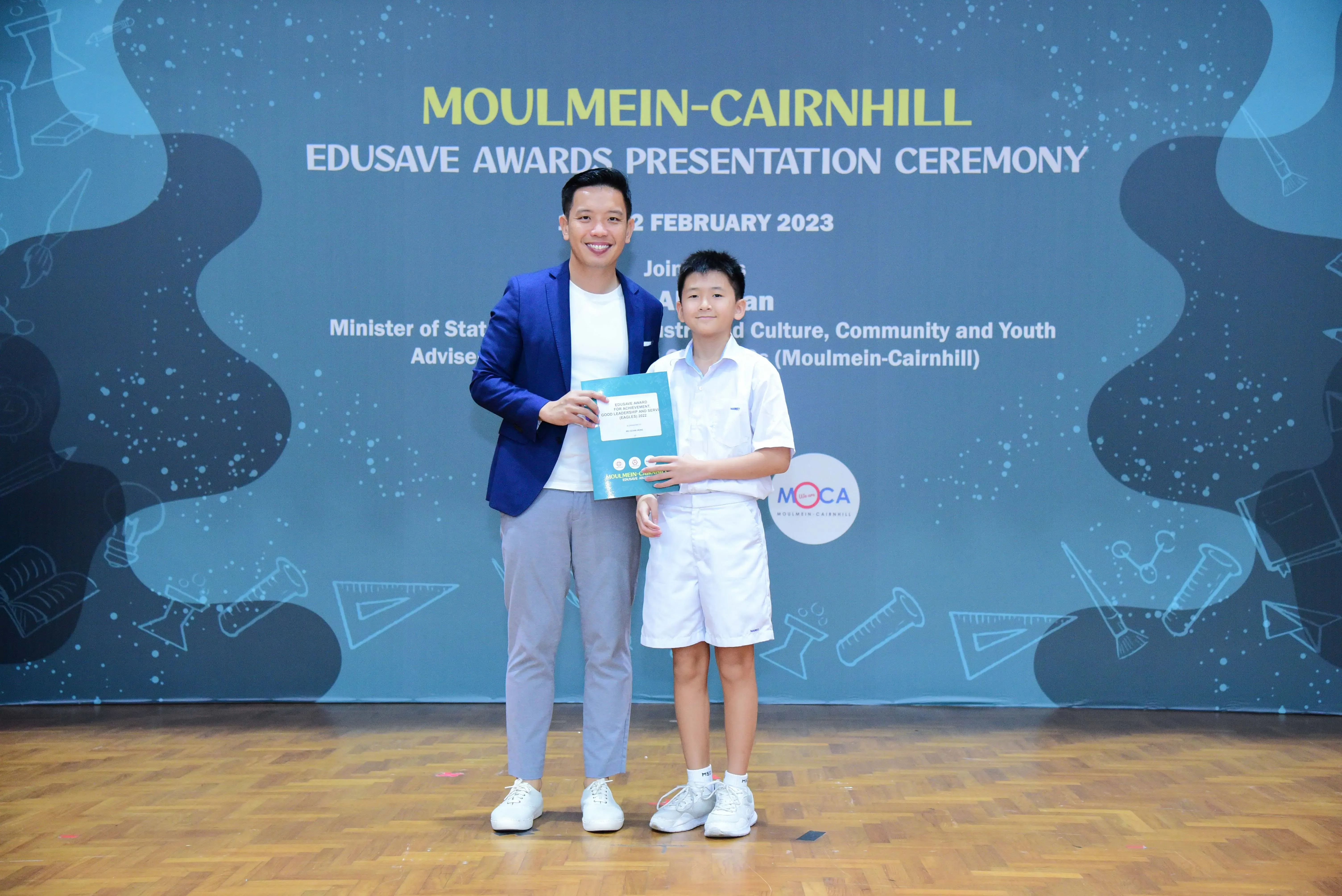 An adult in a blue blazer and a young boy in white uniform hold a certificate at the Moulmein-Cairnhill Edusave Awards 2023.