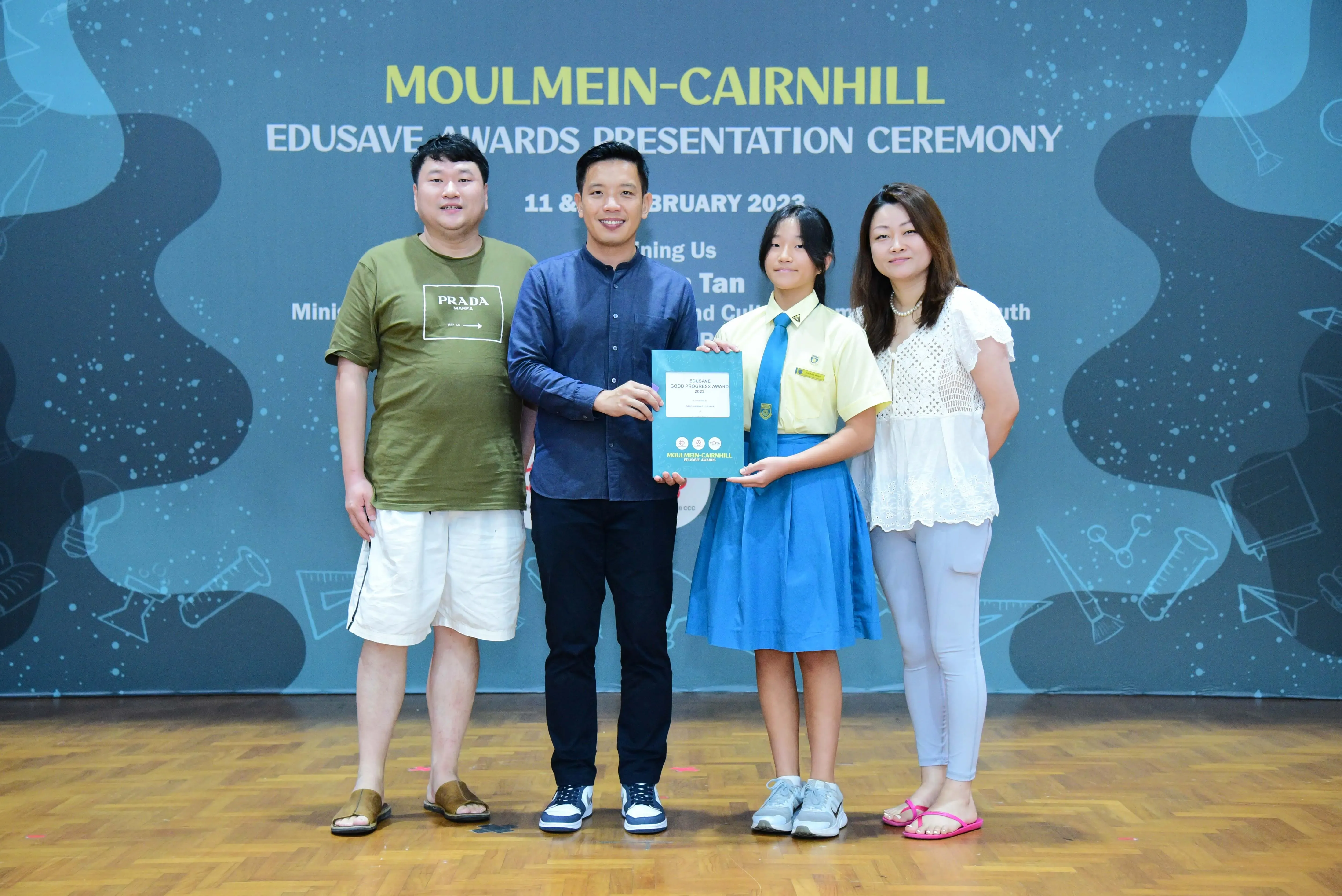 Four people stand before a Moulmein-Cairnhill Edusave Awards Presentation Ceremony banner, with a girl holding an award folder.