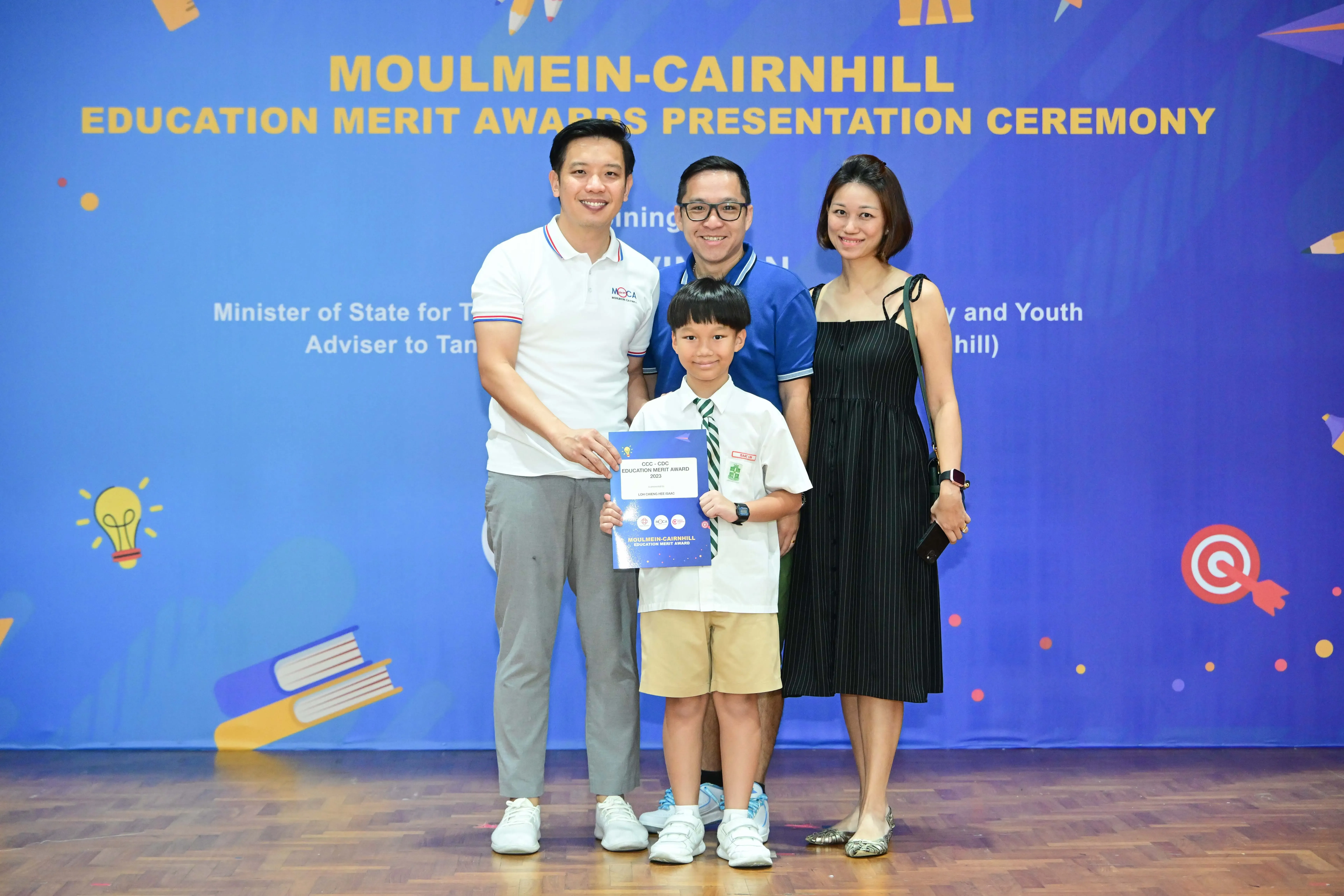 A boy in school uniform holds a certificate with three adults at the Moulmein-Cairnhill Education Merit Awards Ceremony.