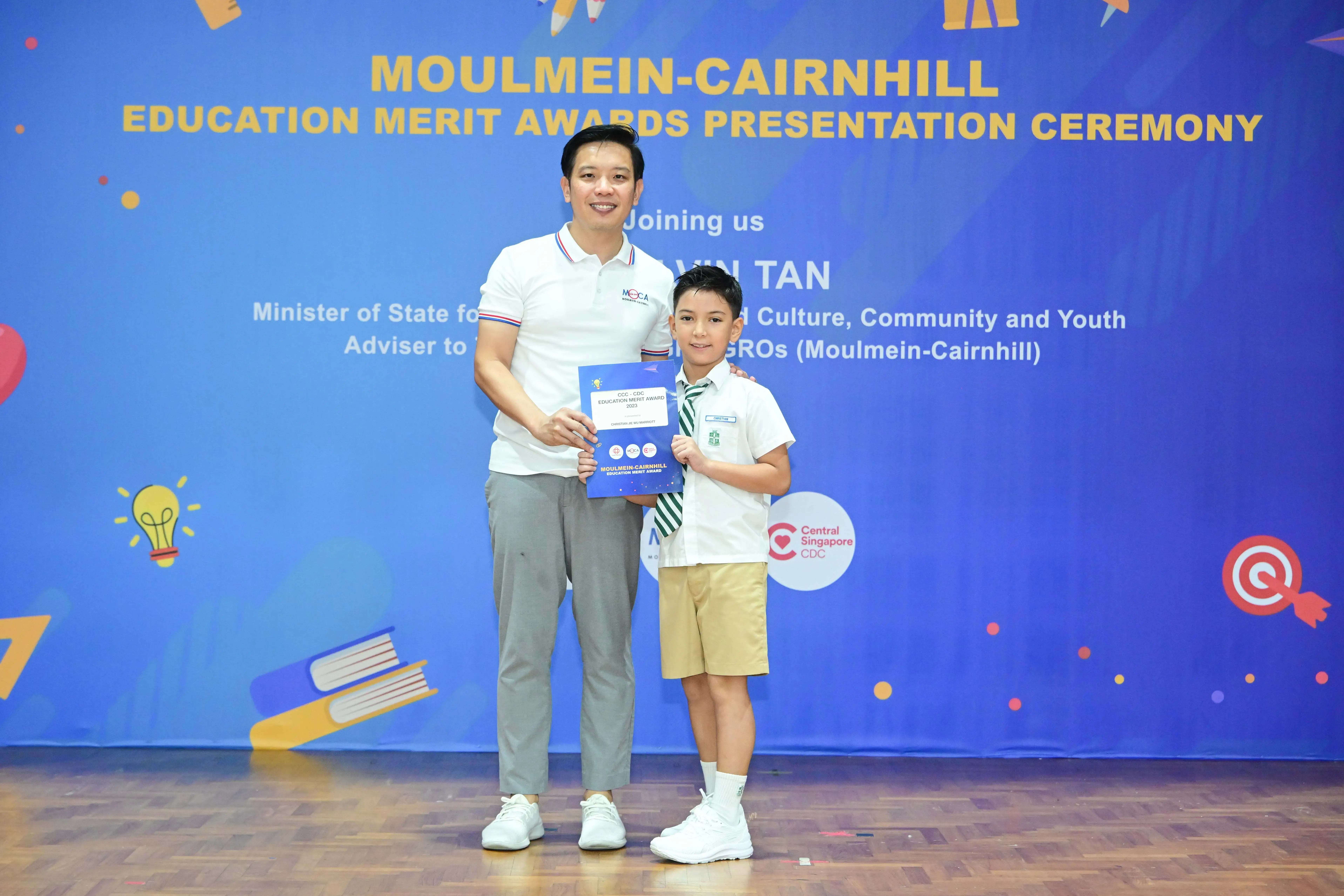 An adult and a young boy hold a certificate at the Moulmein-Cairnhill Education Merit Awards Presentation Ceremony.