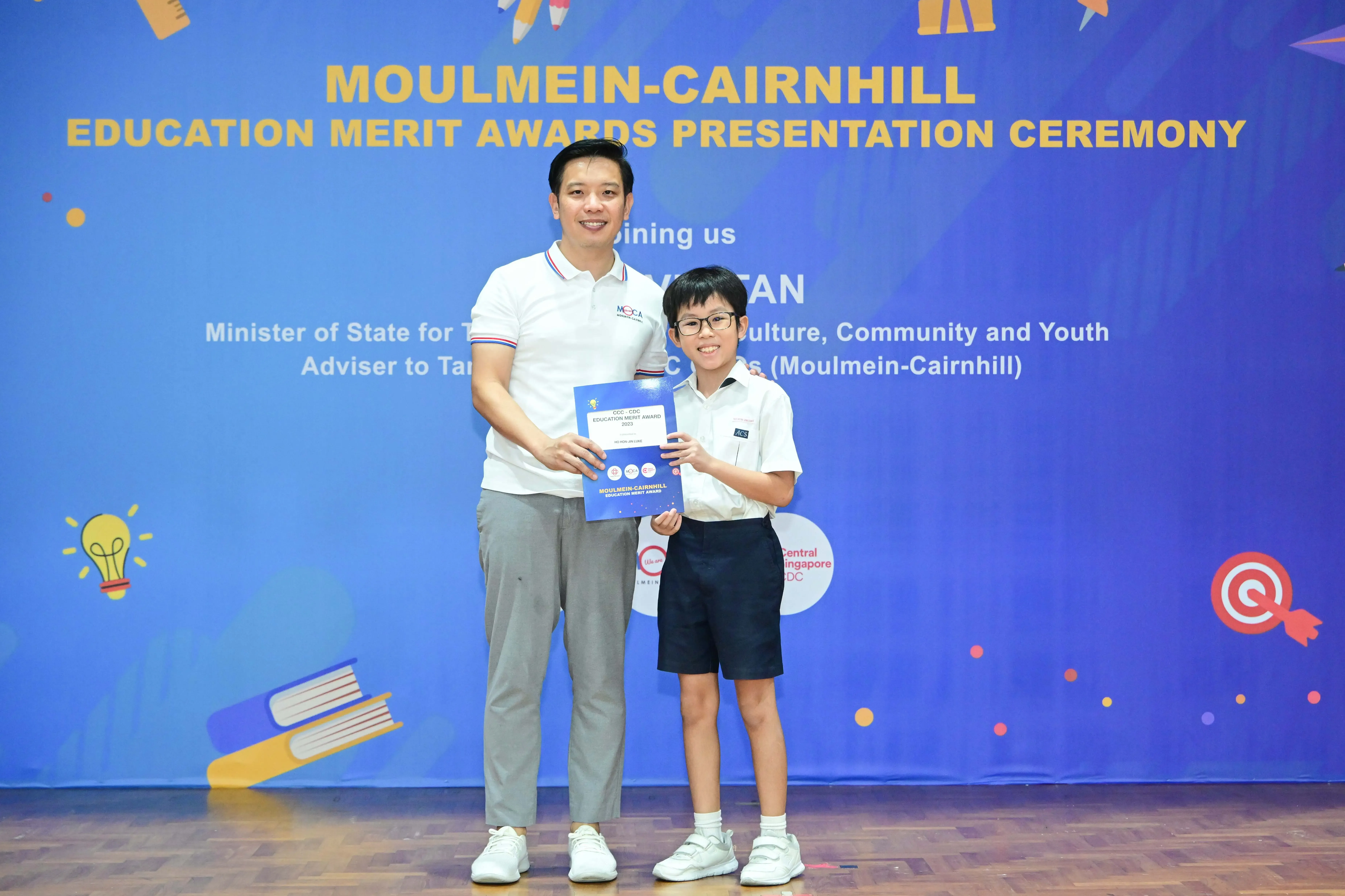 An adult and a young boy hold a certificate together at the Moulmein-Cairnhill Education Merit Awards Presentation Ceremony.