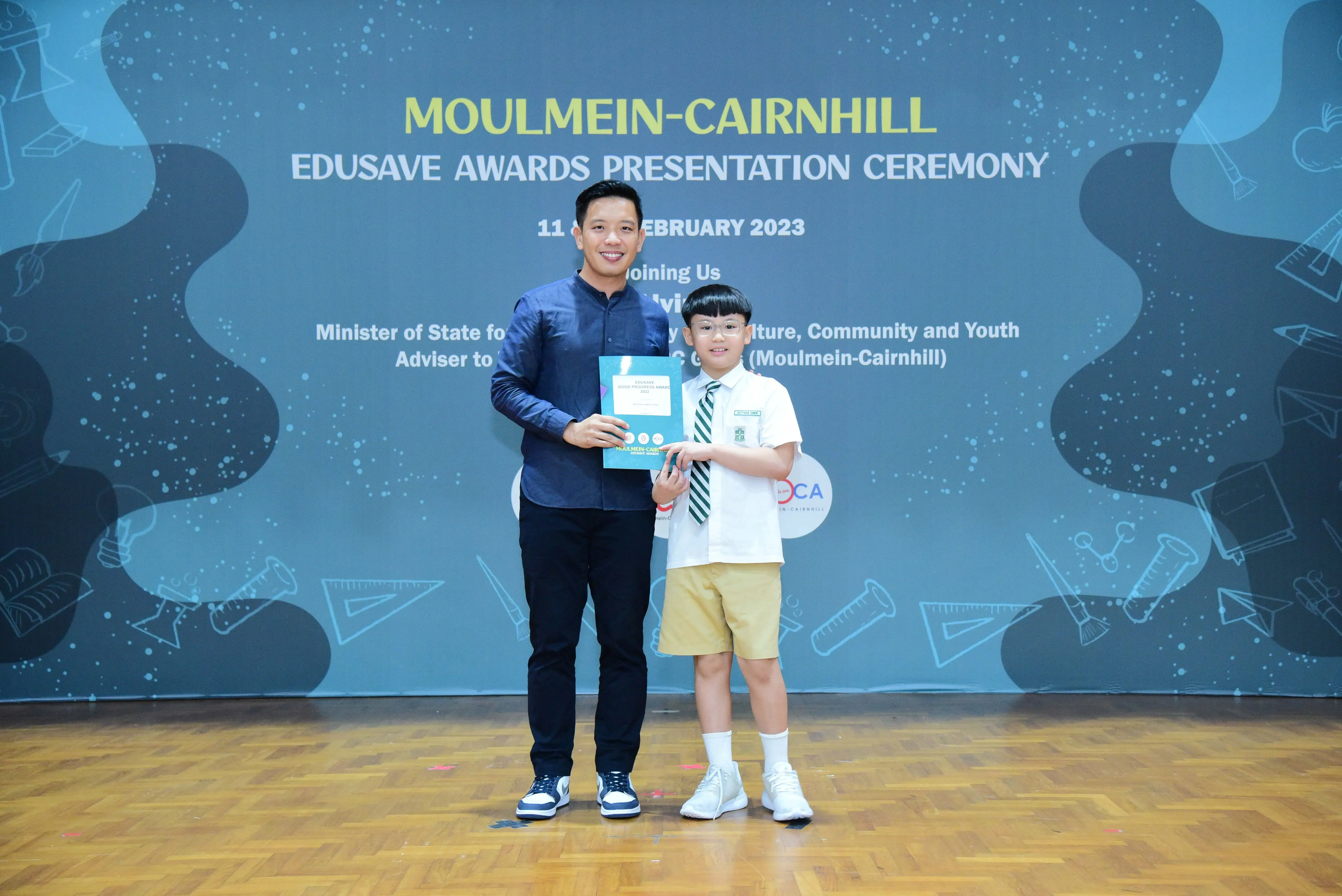 An adult and a young student hold a certificate at the Moulmein-Cairnhill Edusave Awards Presentation Ceremony 2023.
