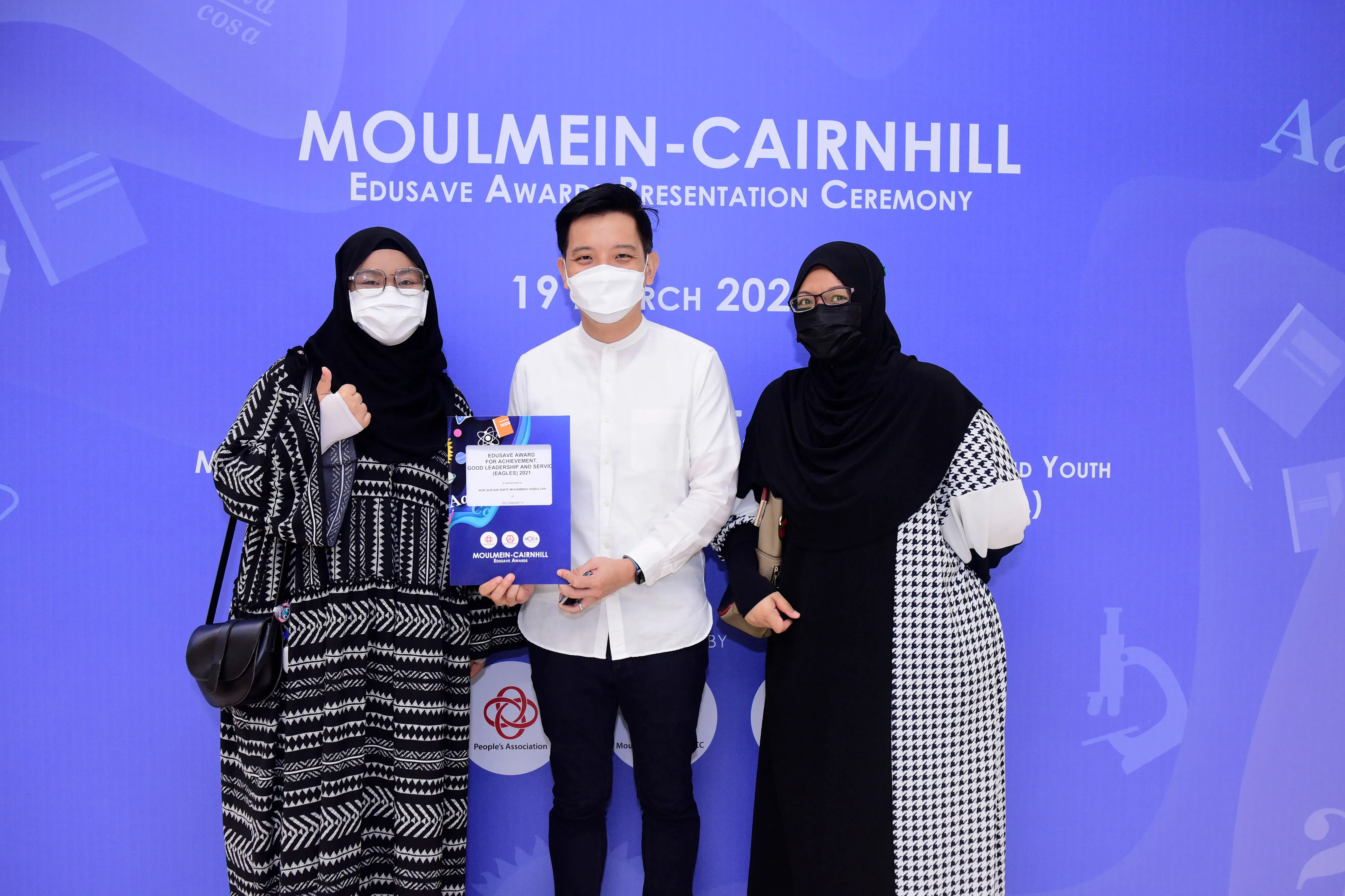 Three masked people pose holding a certificate at the Moulmein-Cairnhill Edusave Award Presentation Ceremony backdrop.