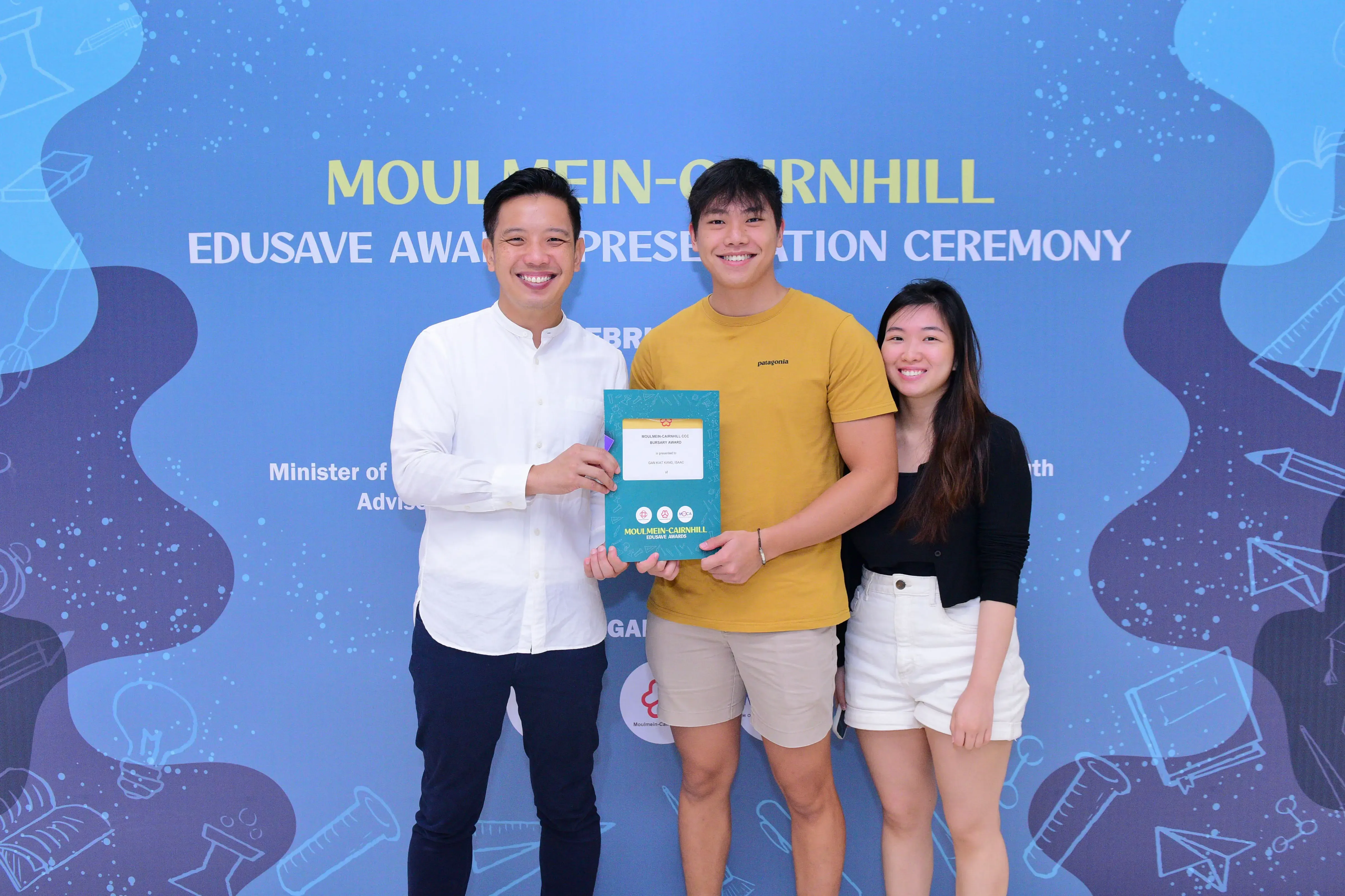 Three smiling people pose together holding a certificate at the Moulmein-Cairnhill Edusave Awards Presentation Ceremony.