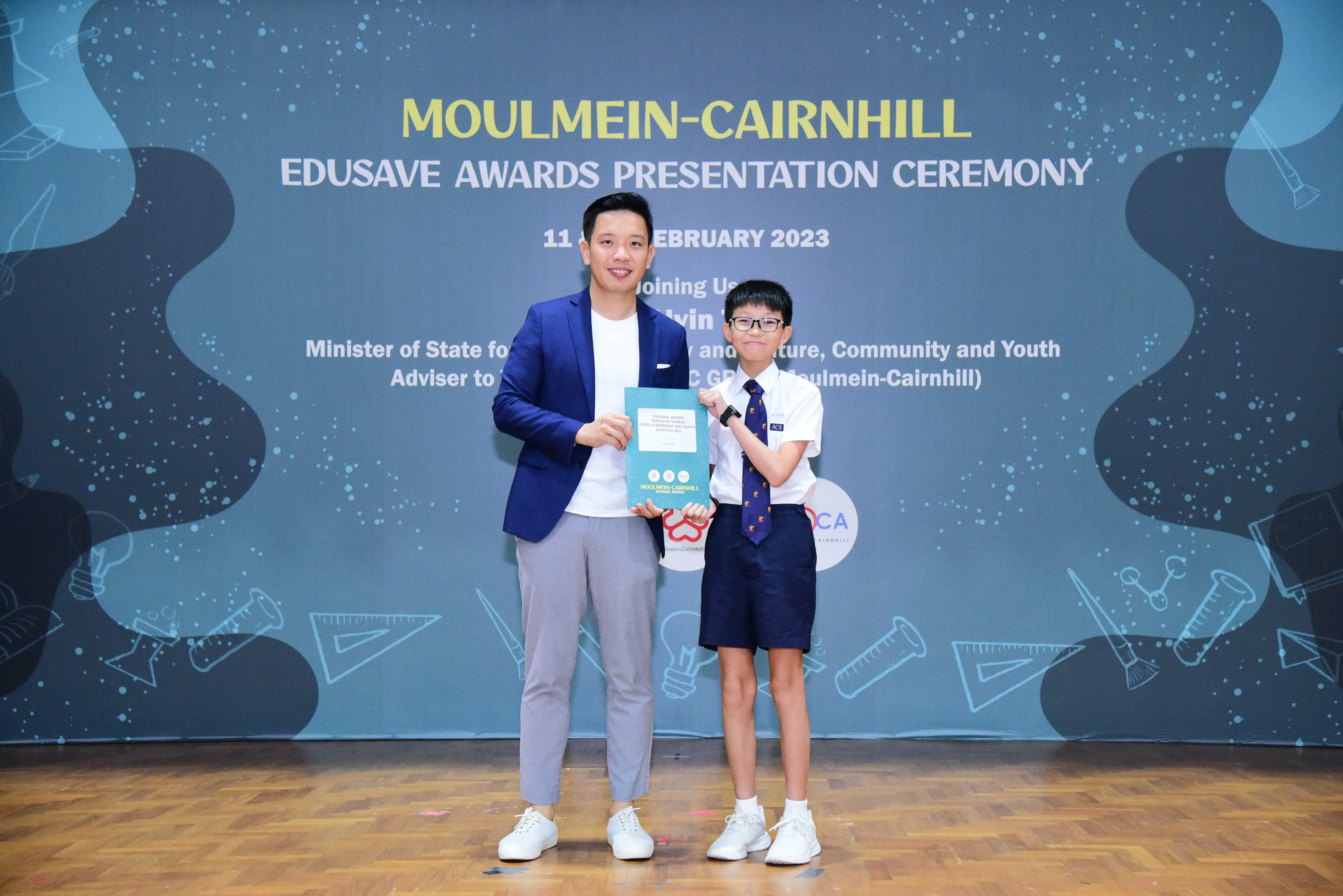 An adult in a blue blazer and a young student in uniform hold a certificate at the Moulmein-Cairnhill Edusave Awards Ceremony 2023.