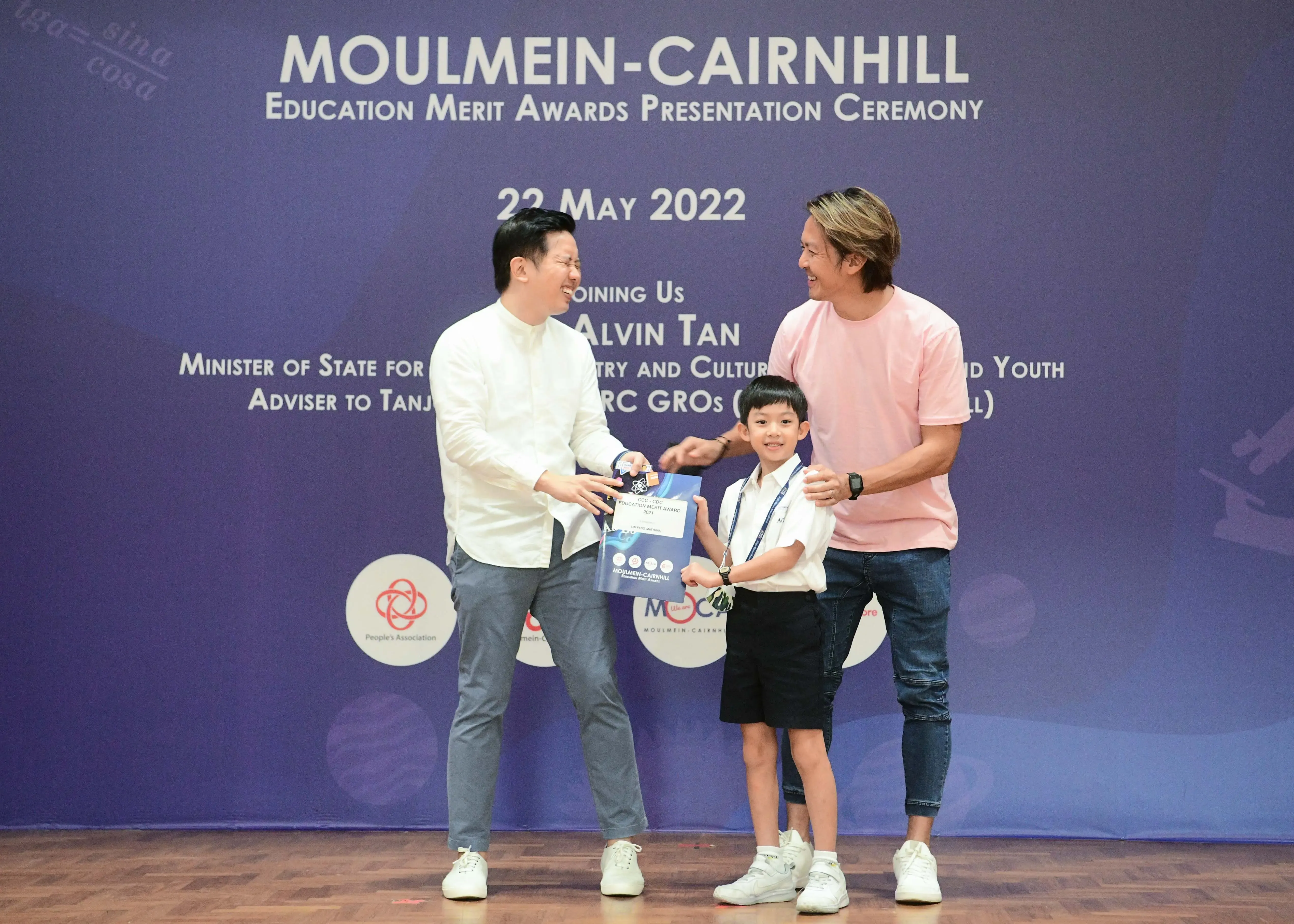 Two men and a young boy in school uniform hold an award folder on stage at the Moulmein-Cairnhill Education Merit Awards 2022.