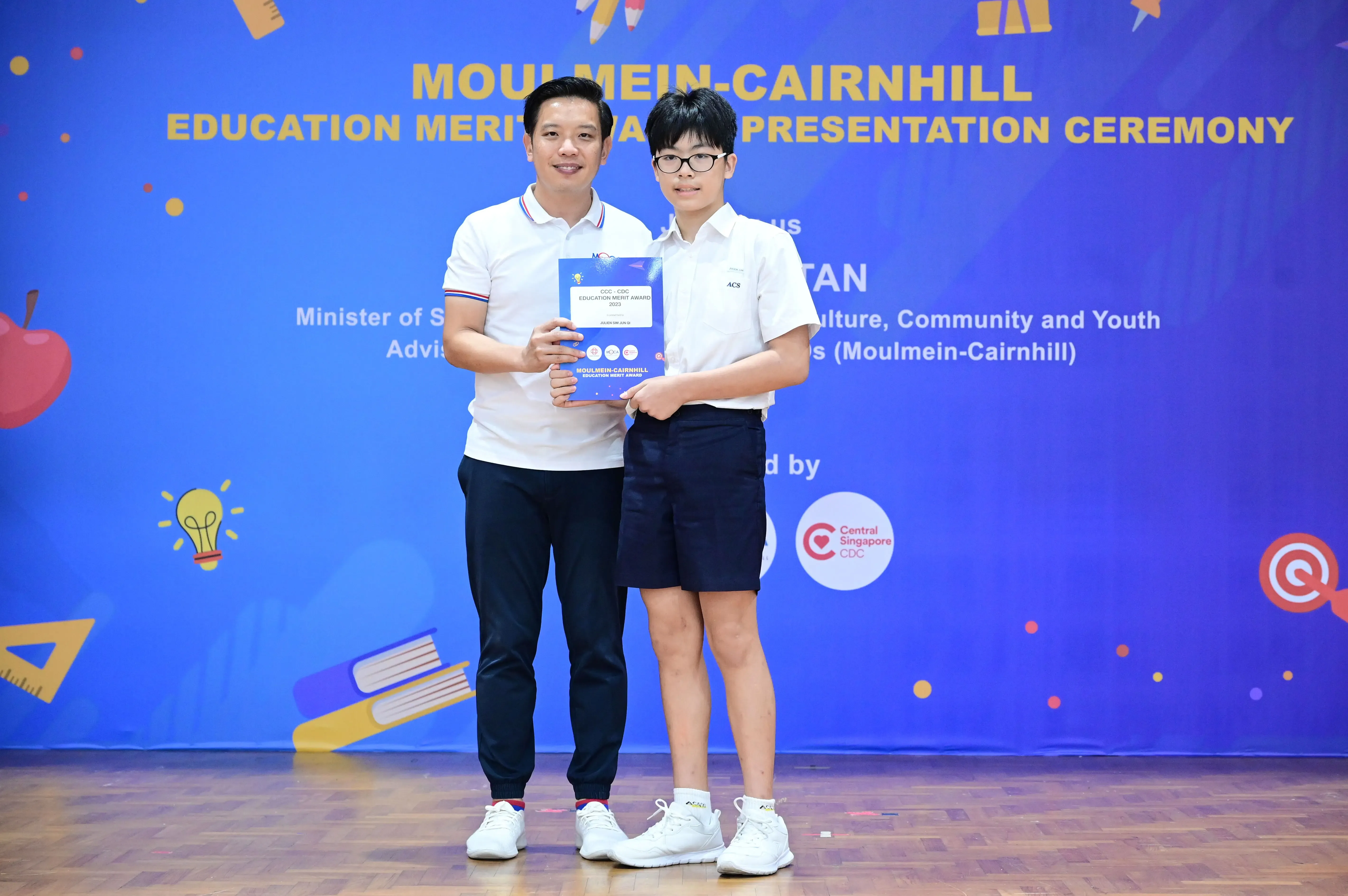 An adult and a student in school uniform hold a certificate at the Moulmein-Cairnhill Education Merit Award ceremony.