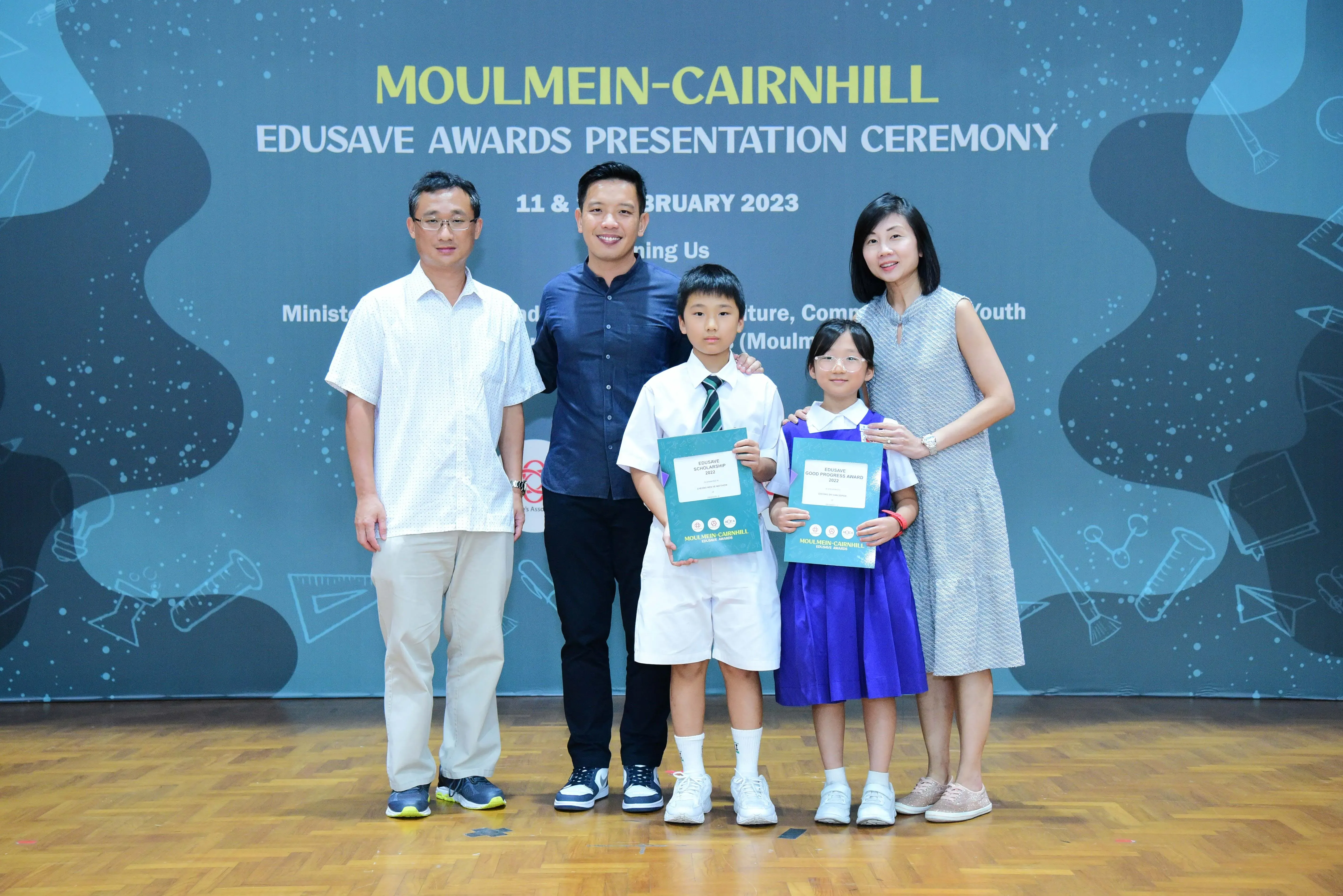 Five people pose at the Moulmein-Cairnhill Edusave Awards Ceremony; two children hold teal award folders.