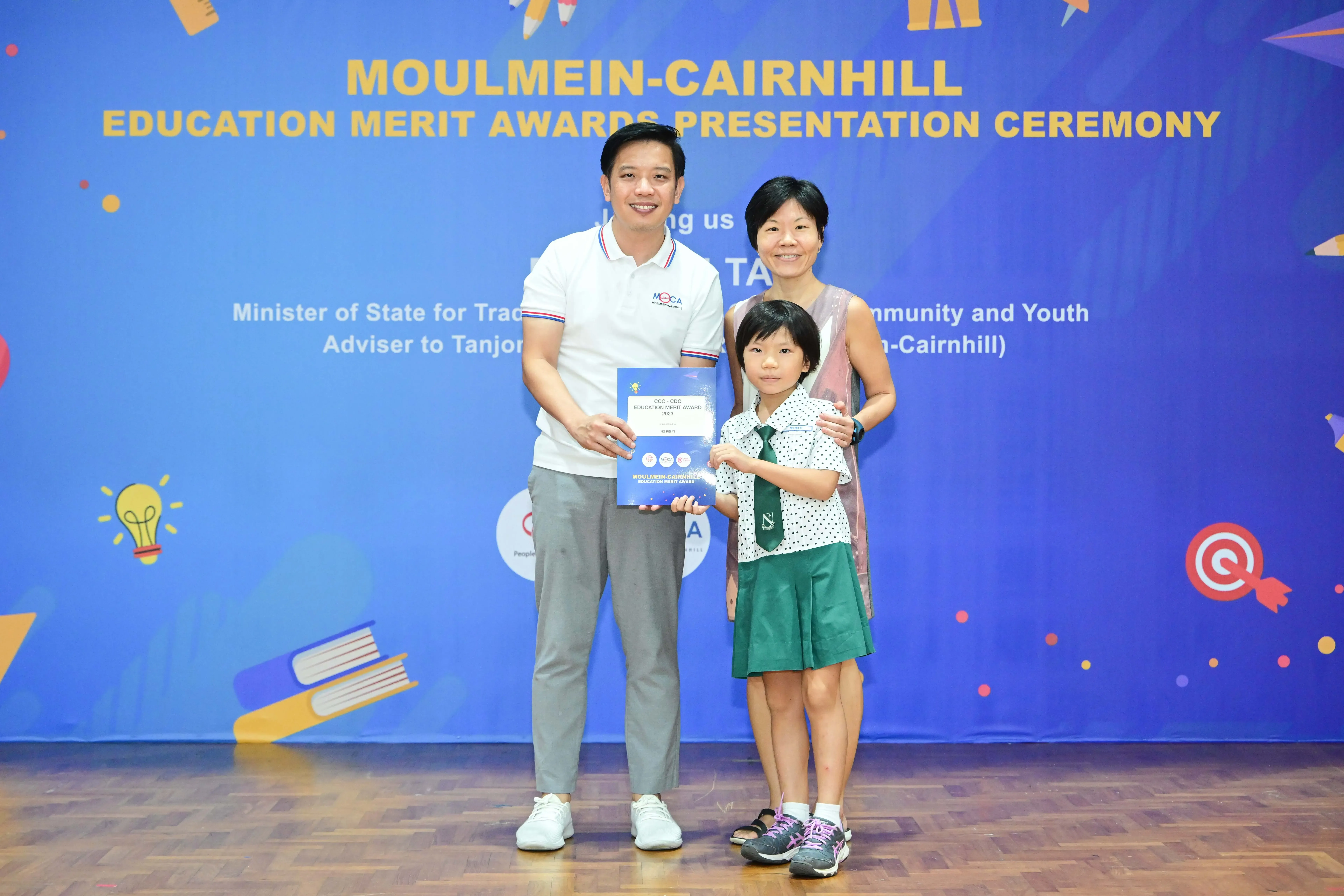 A man, woman, and young girl in school uniform hold an award certificate at the Moulmein-Cairnhill Education Merit Awards ceremony.