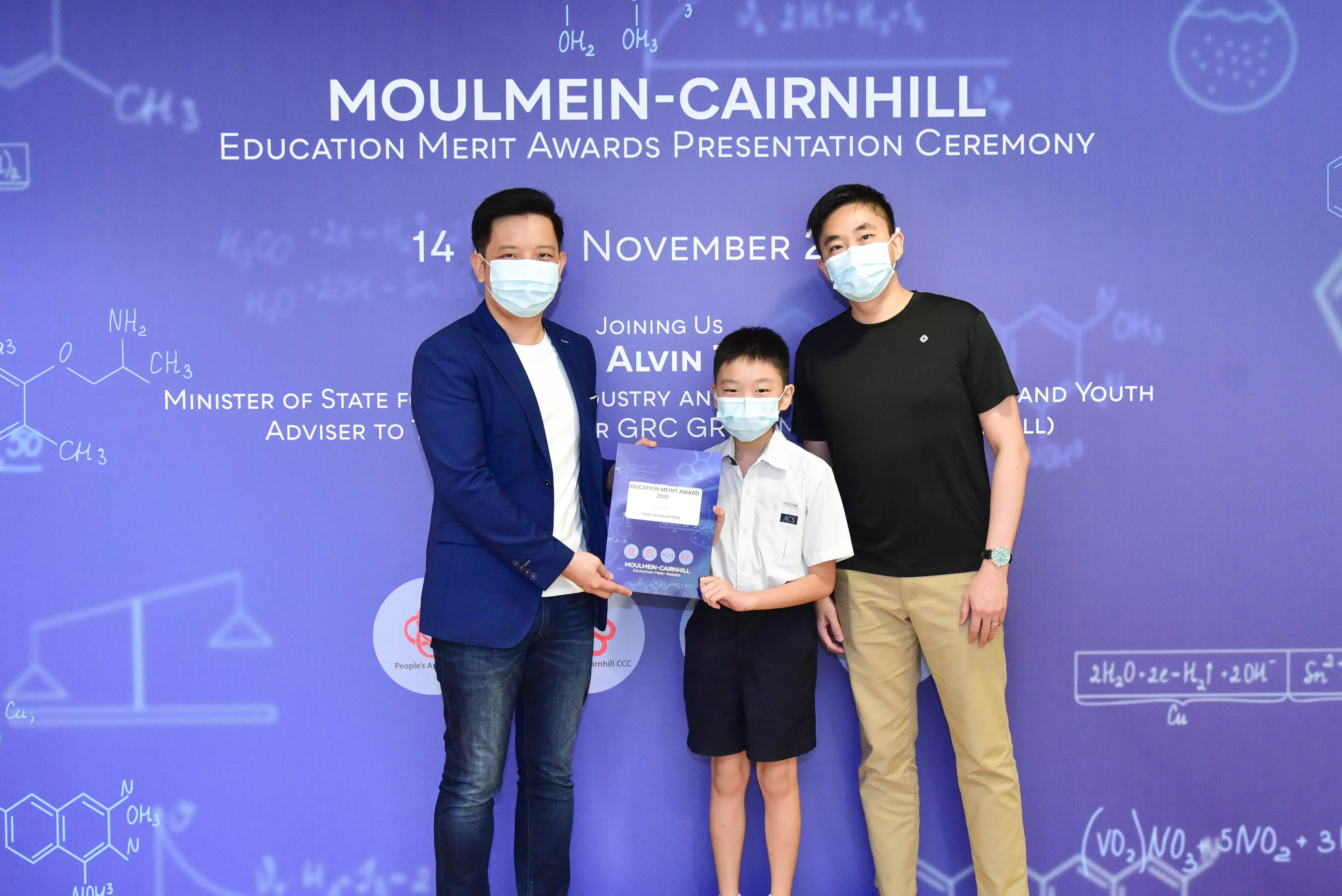 Two masked men and a masked boy hold an award certificate at the Moulmein-Cairnhill Education Merit Awards ceremony.