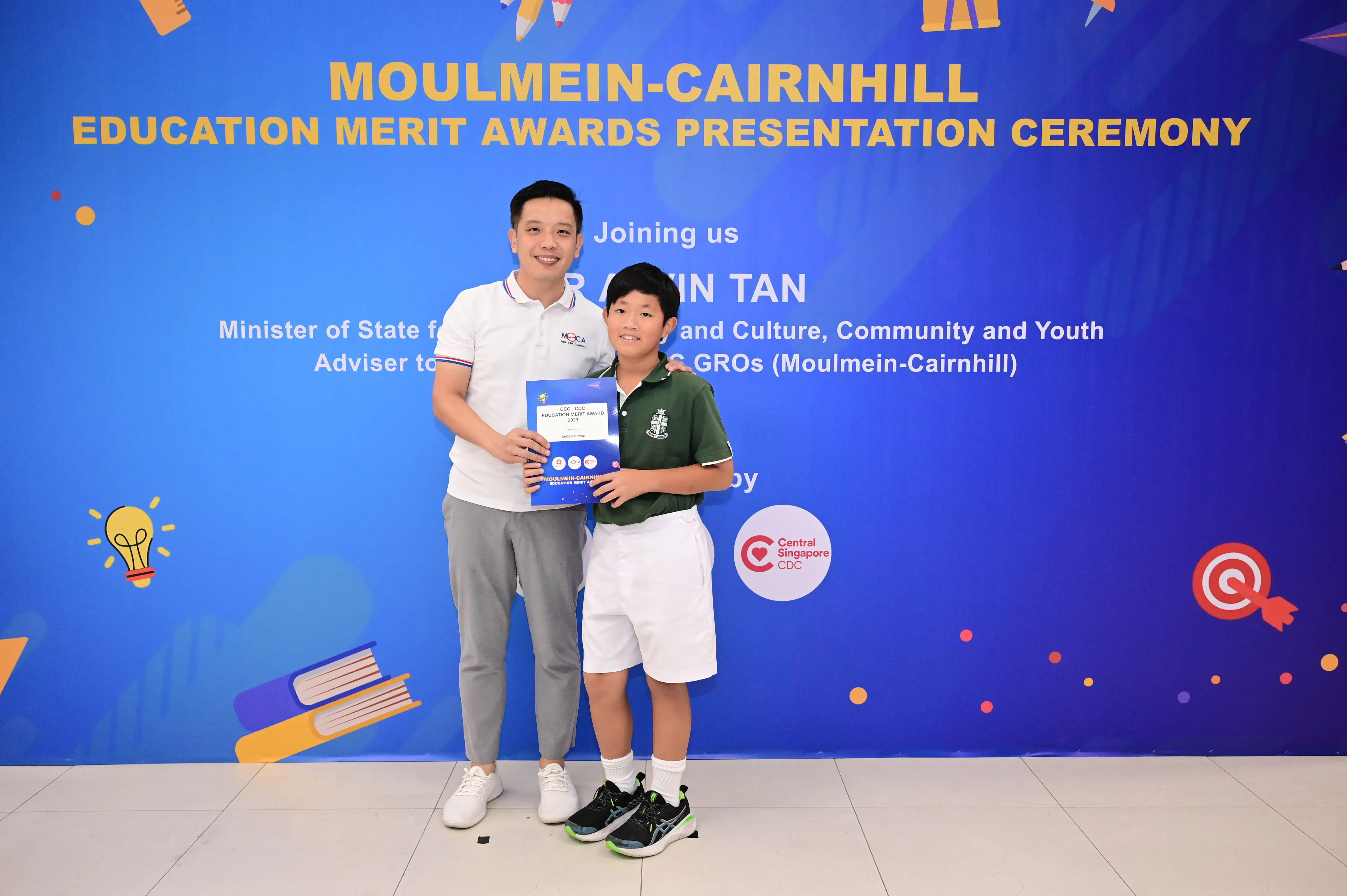 A man and a young boy hold a blue award booklet at the Moulmein-Cairnhill Education Merit Awards Ceremony.