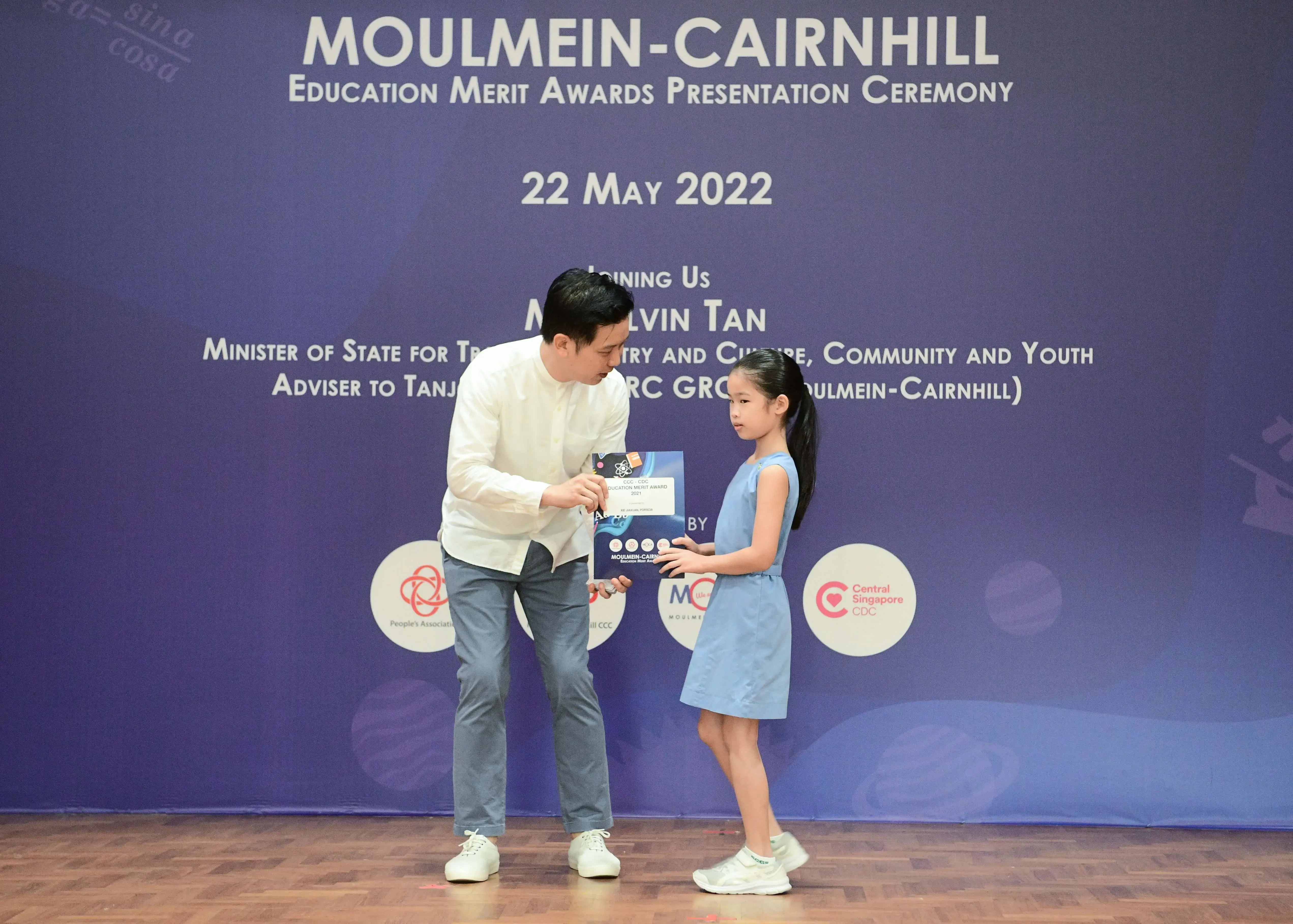 A man presents an award certificate to a young girl on stage at the Moulmein-Cairnhill Education Merit Awards ceremony, May 2022.