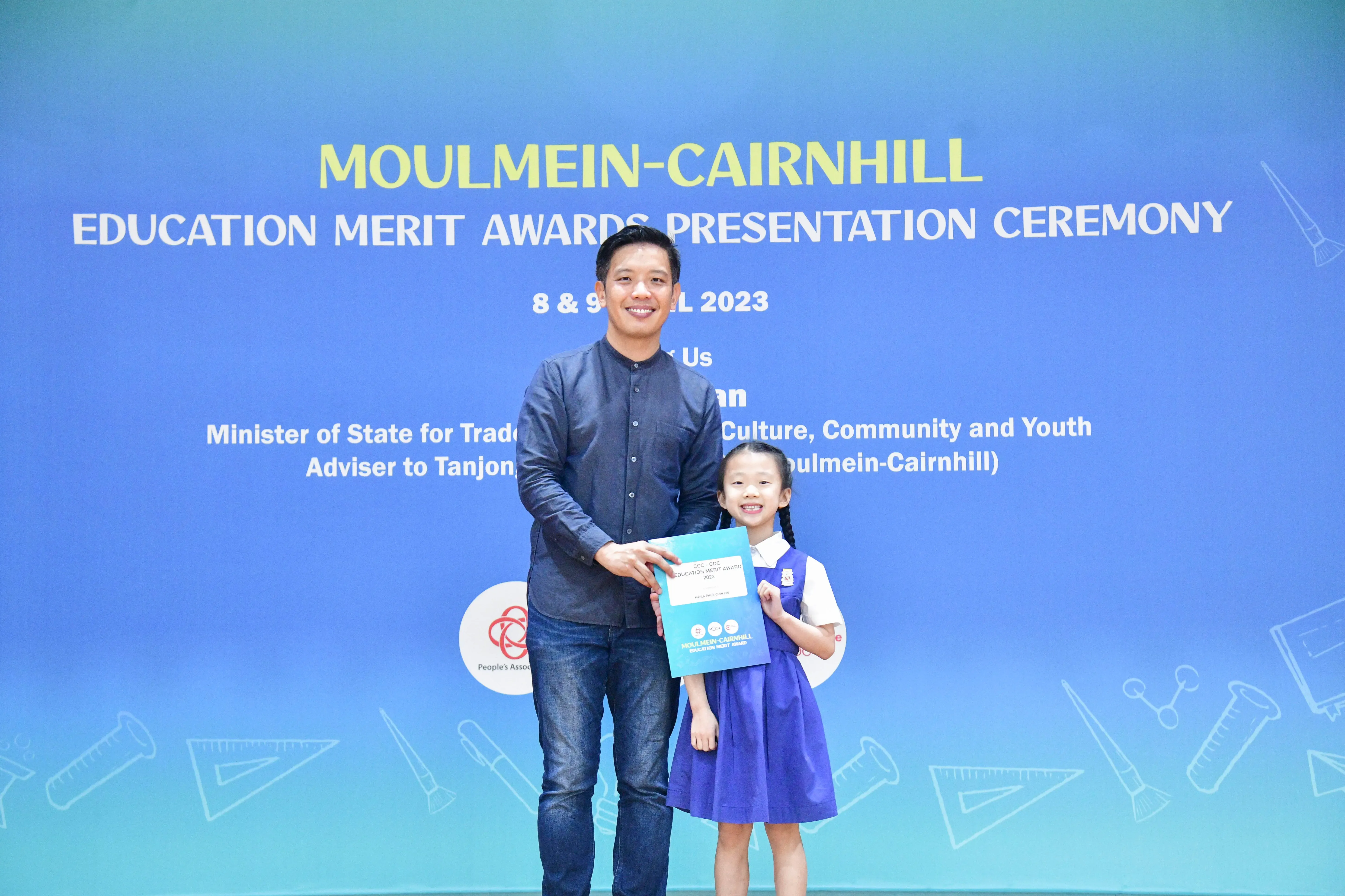 A man and a young girl hold a certificate together in front of a Moulmein-Cairnhill Education Merit Awards banner.