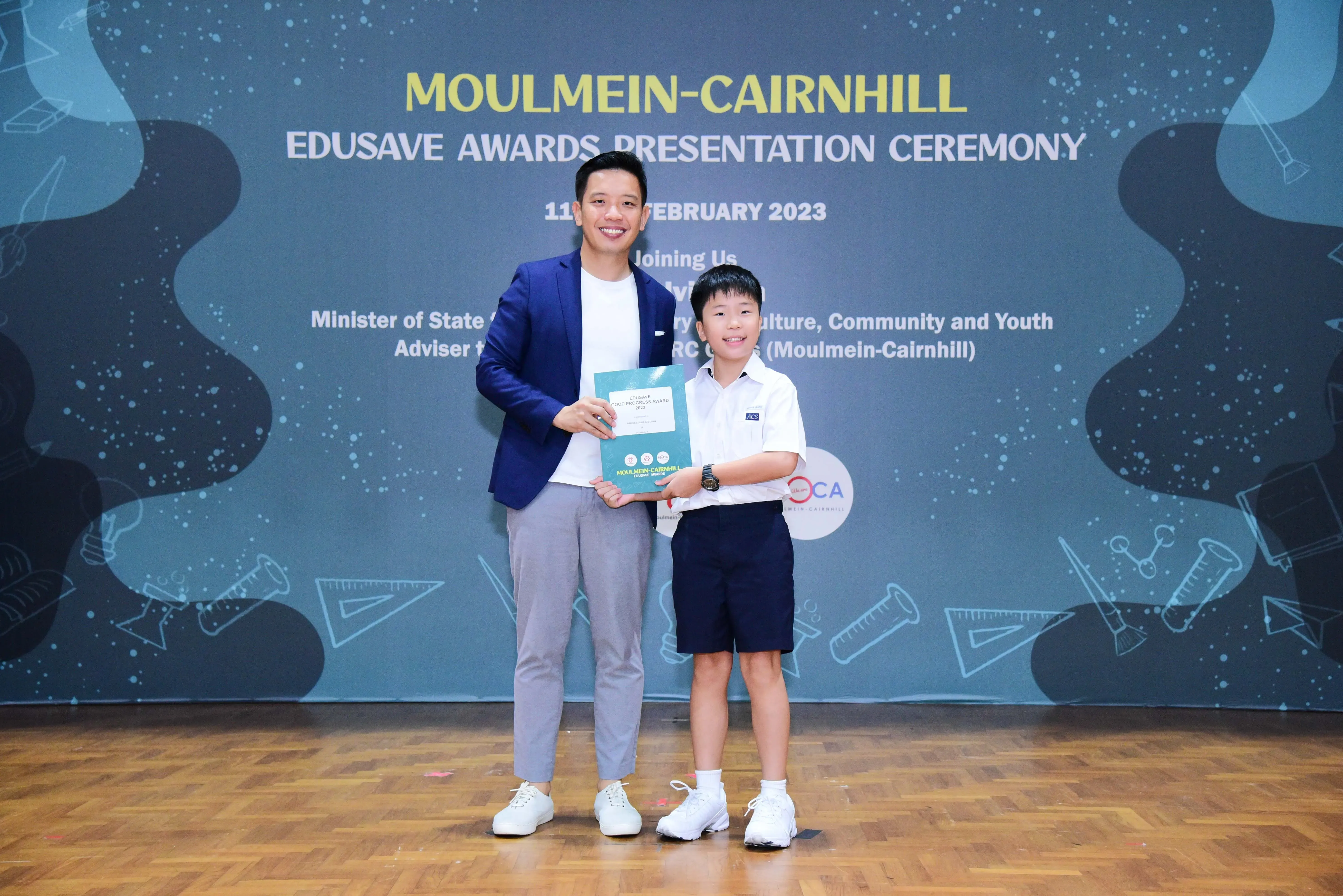 A man in a blue blazer and a young boy in school uniform hold a certificate at the Moulmein-Cairnhill Edusave Awards 2023.