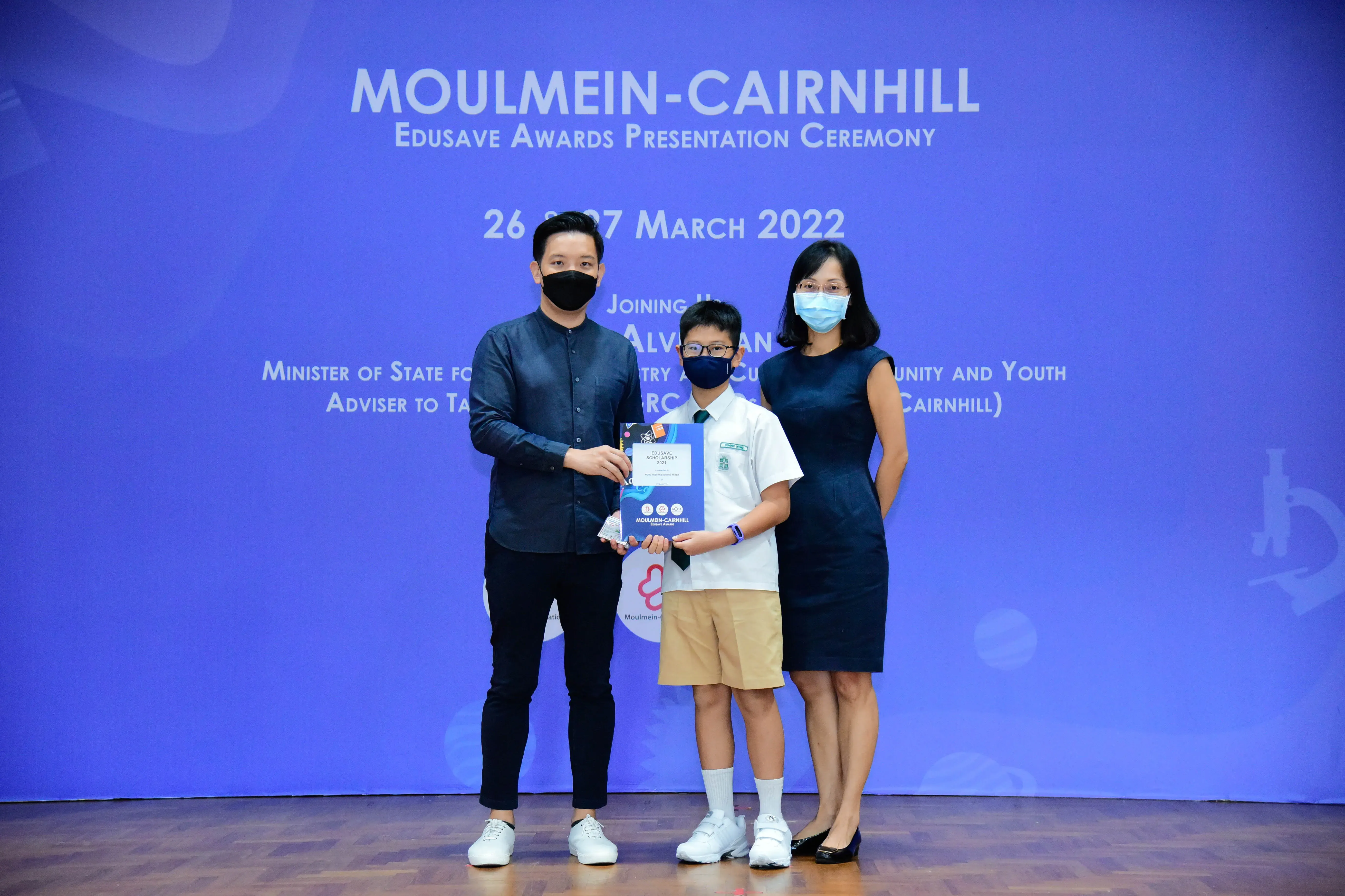 Three masked individuals pose at the Moulmein-Cairnhill Edusave Awards Ceremony on 26-27 March 2022, holding a certificate.