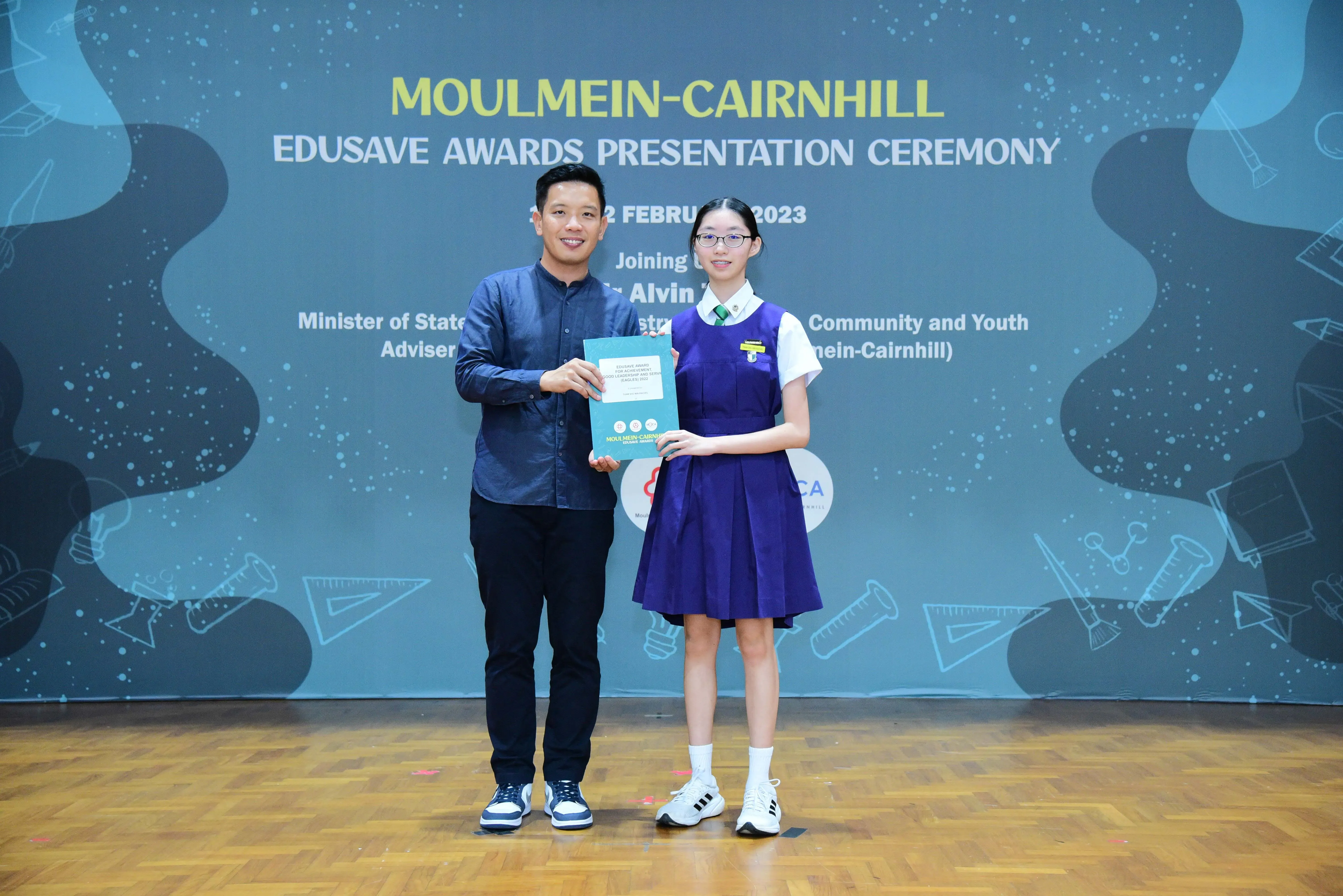 A man and a girl in school uniform hold a certificate together at the Moulmein-Cairnhill Edusave Awards Ceremony 2023.