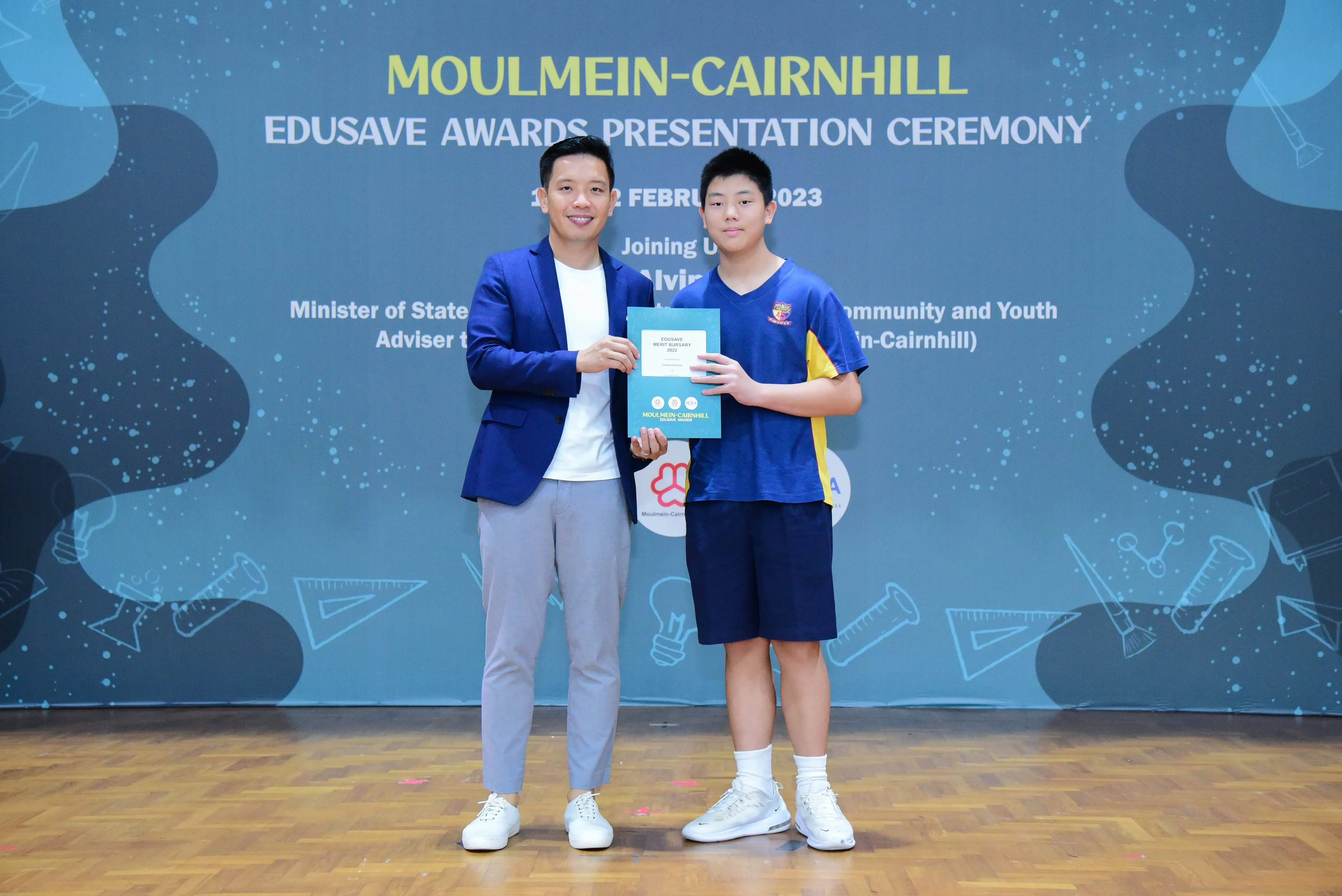 A man in a blue blazer and a young student hold a certificate at the Moulmein-Cairnhill Edusave Awards Ceremony 2023.