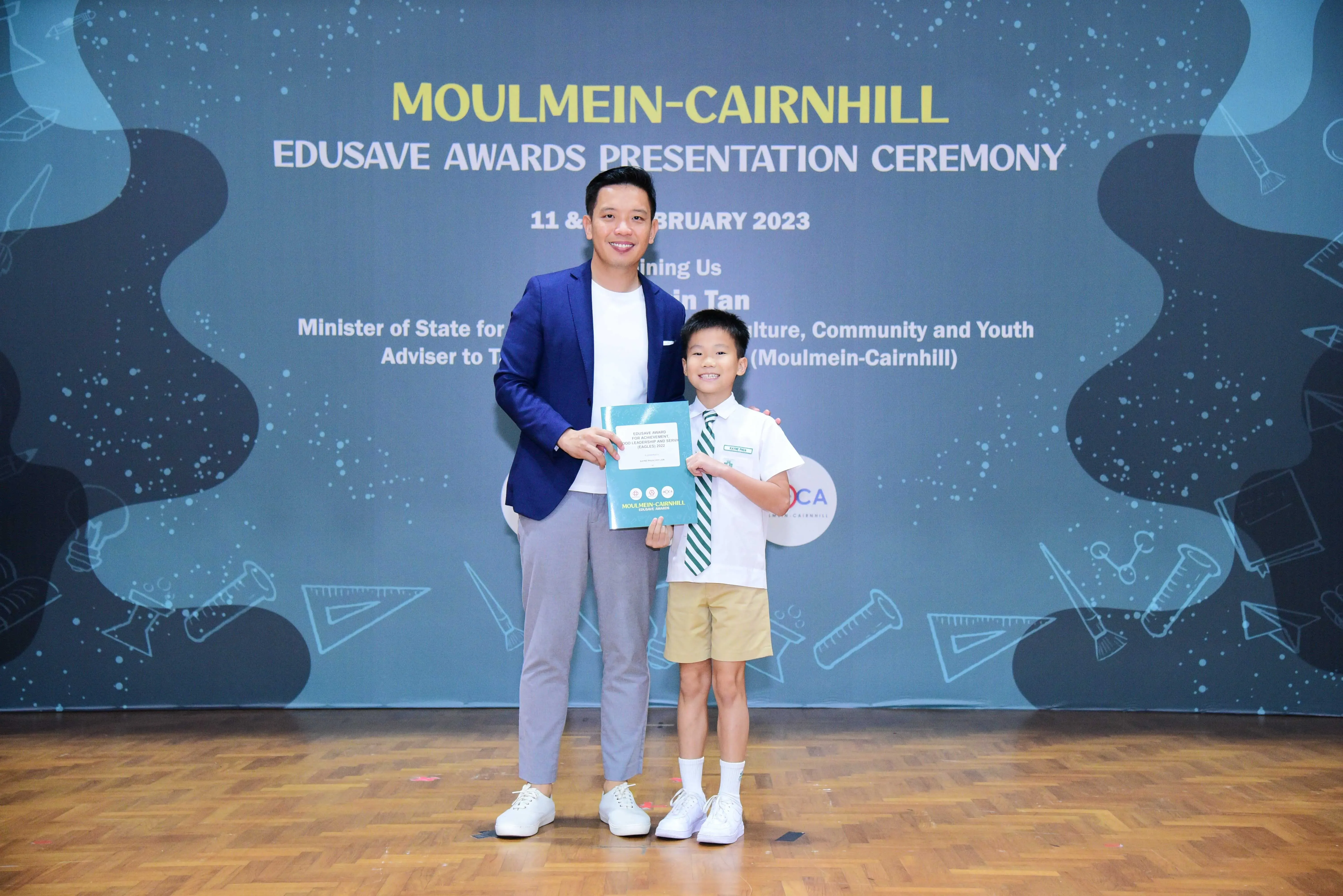 A man in a blue blazer and a young boy in school uniform hold a certificate at the Moulmein-Cairnhill Edusave Awards 2023.