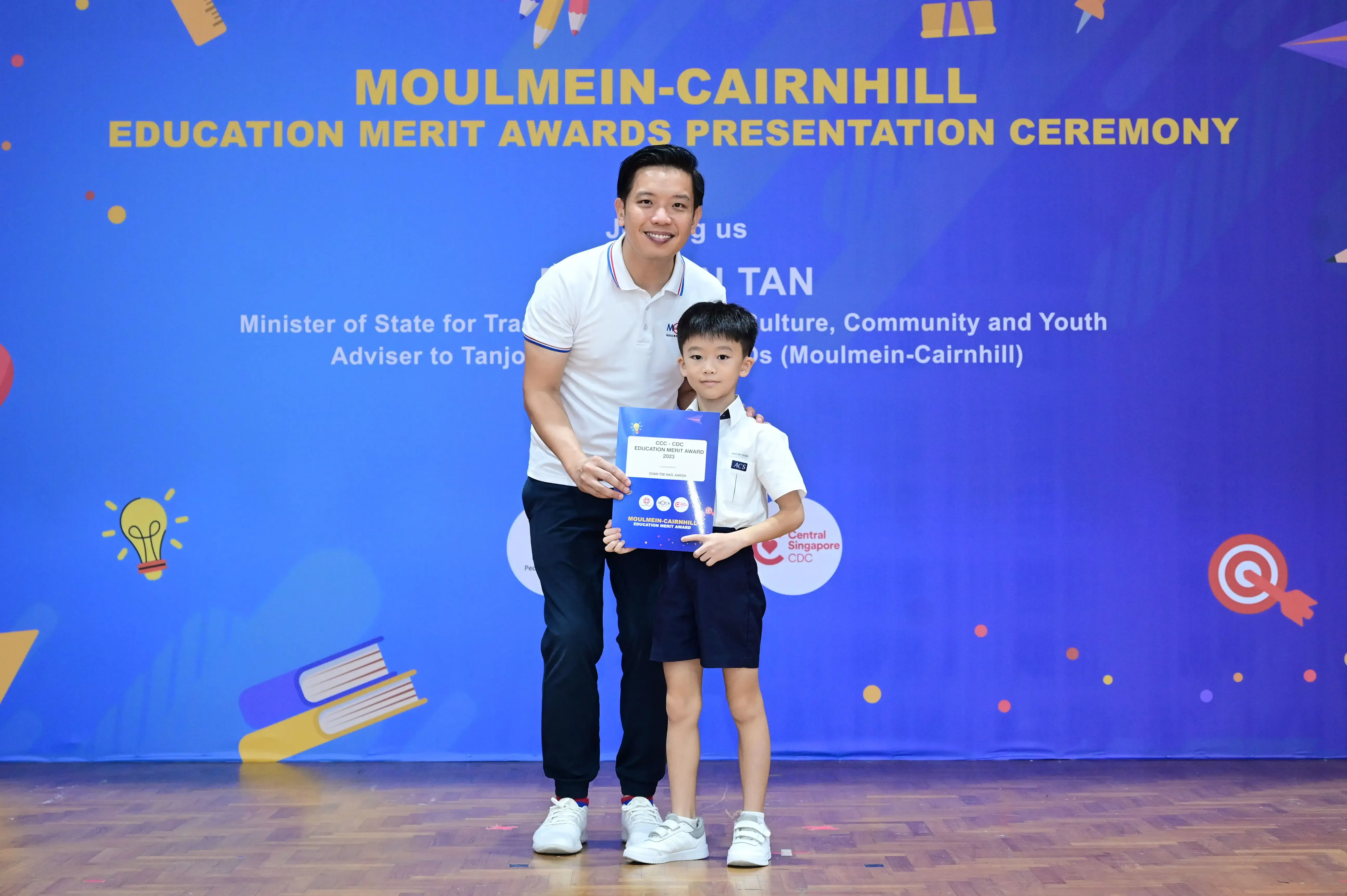 A man and young boy hold an award certificate at the Moulmein-Cairnhill Education Merit Awards Presentation Ceremony.
