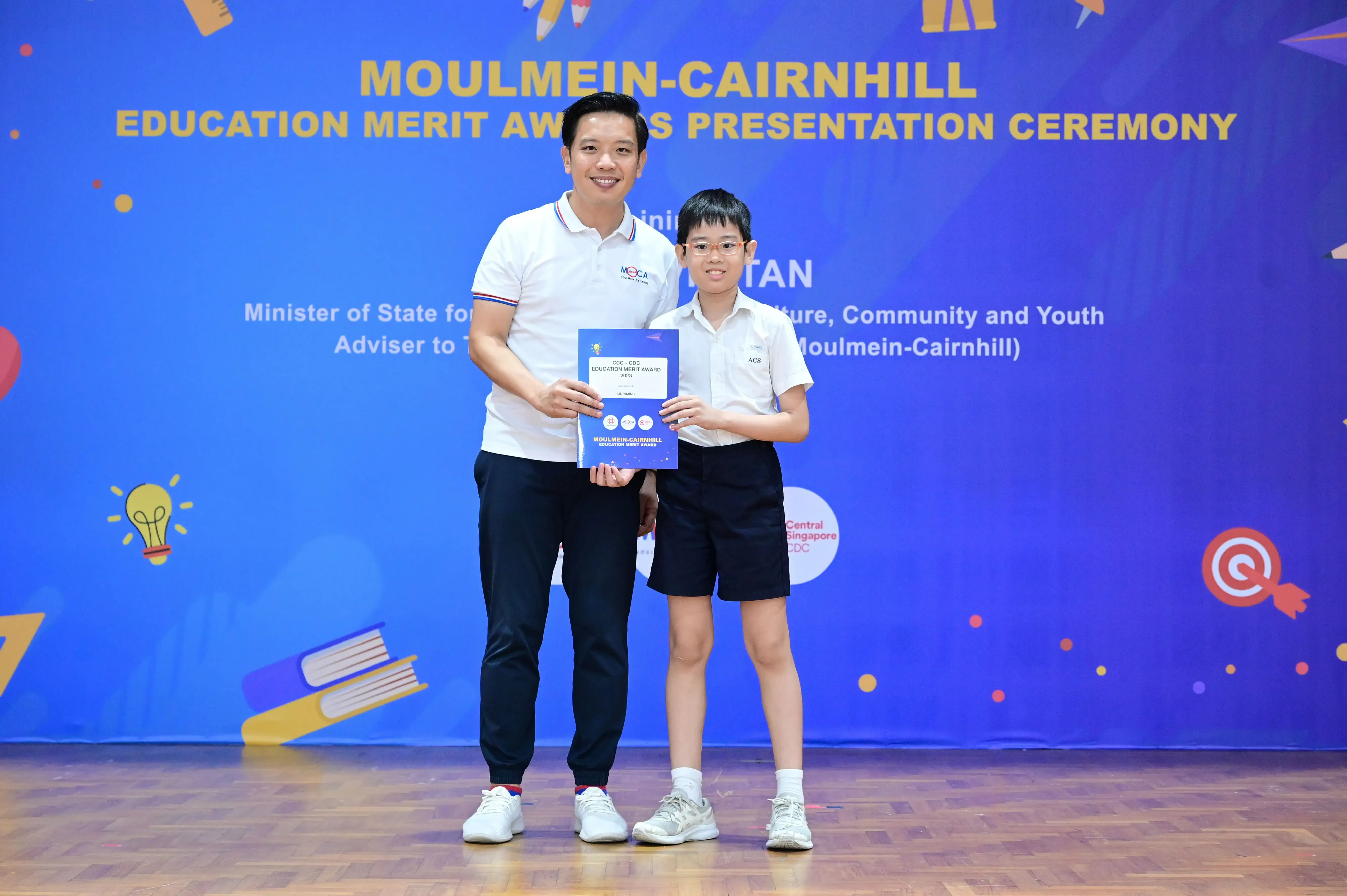 An adult and a young student hold a certificate at the Moulmein-Cairnhill Education Merit Awards Presentation Ceremony.