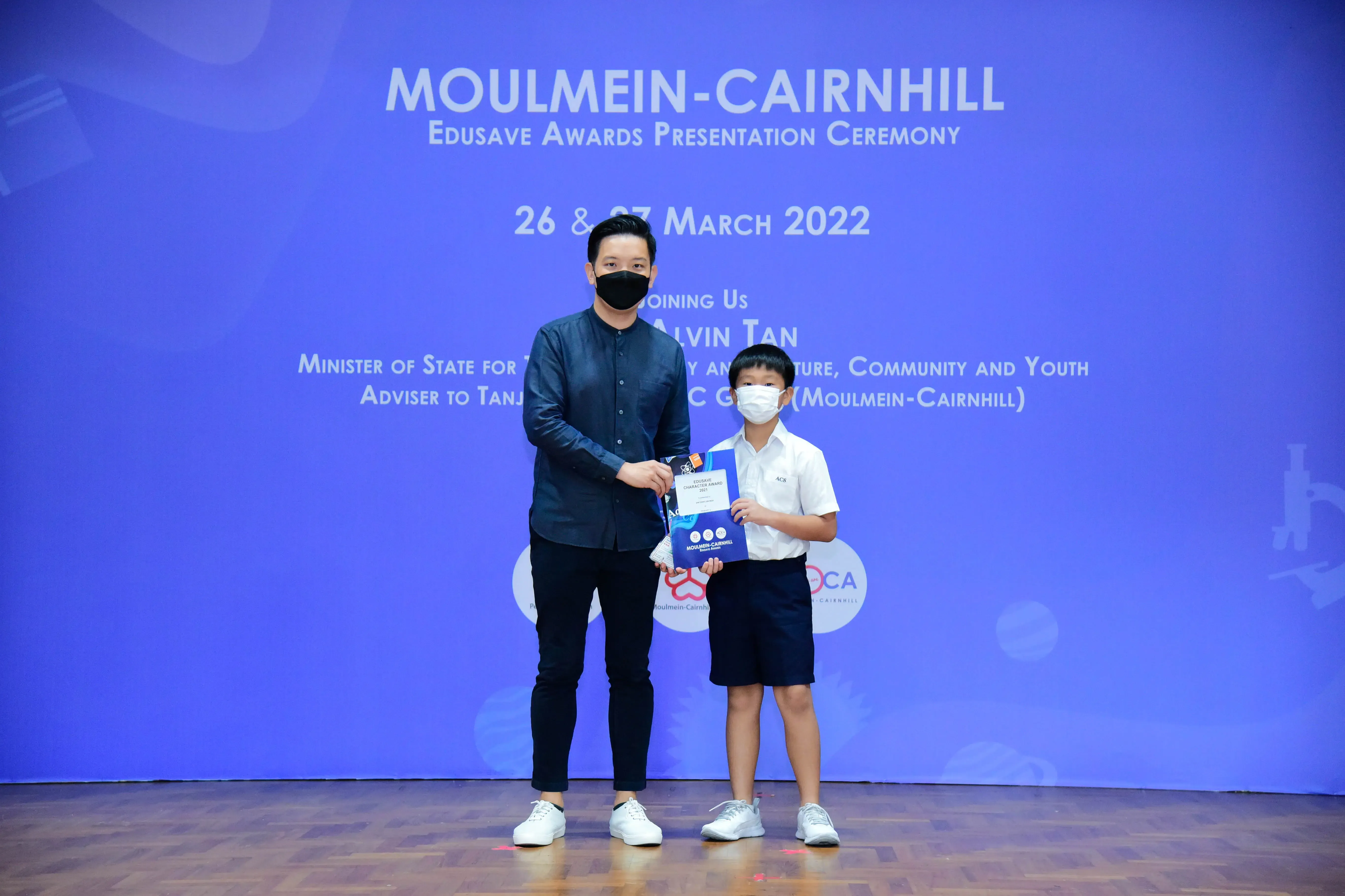 An adult and a young boy in masks hold an award at the Moulmein-Cairnhill Edusave Awards Ceremony, March 2022.