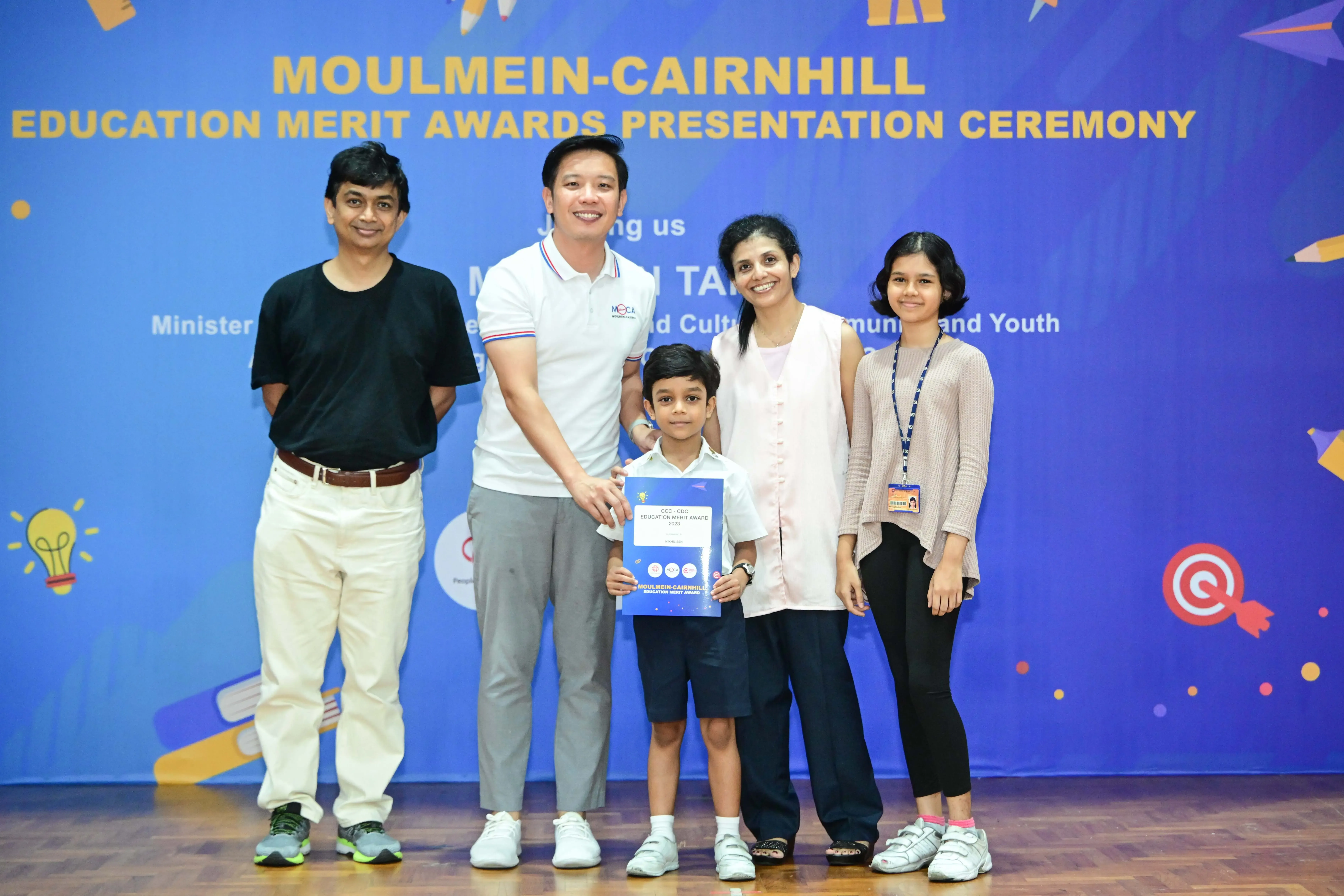 Five people pose before a blue Moulmein-Cairnhill Education Merit Awards Presentation Ceremony banner, a boy holds a certificate.