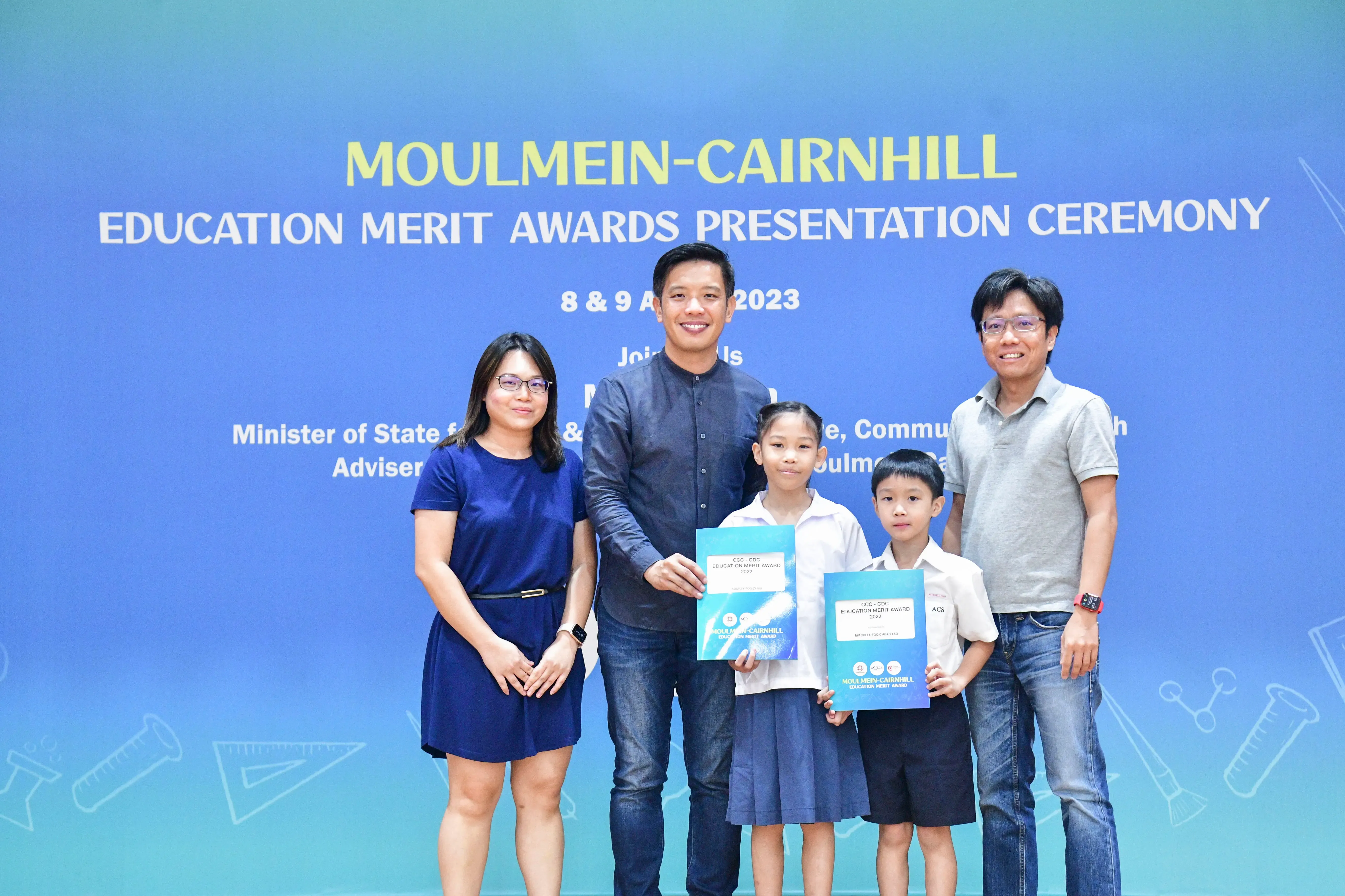 Five people pose before a Moulmein-Cairnhill Education Merit Awards Presentation Ceremony banner, two children holding award folders.