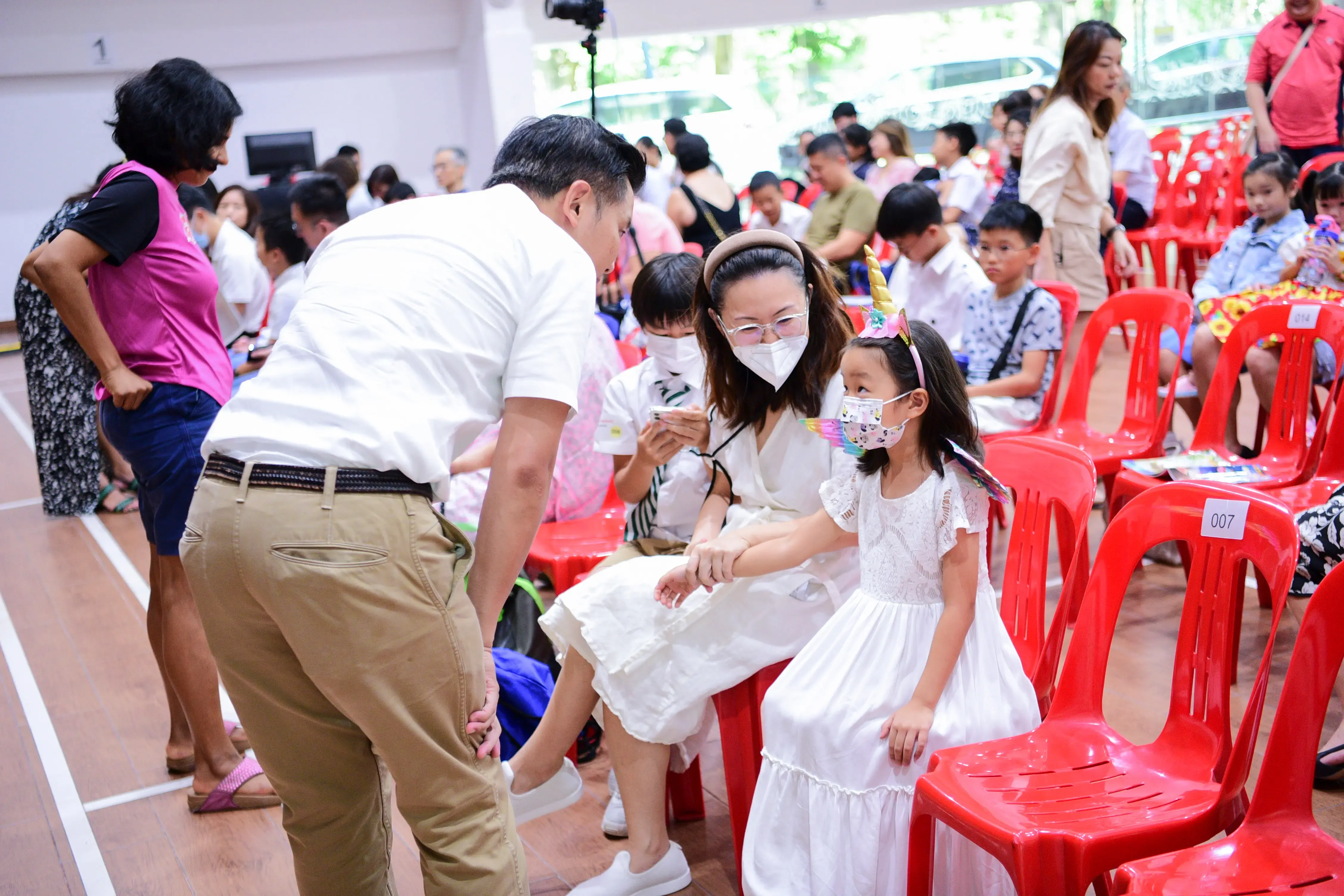 Adults and masked children sit on red chairs in a crowded hall; a man leans toward a girl in a unicorn headband.