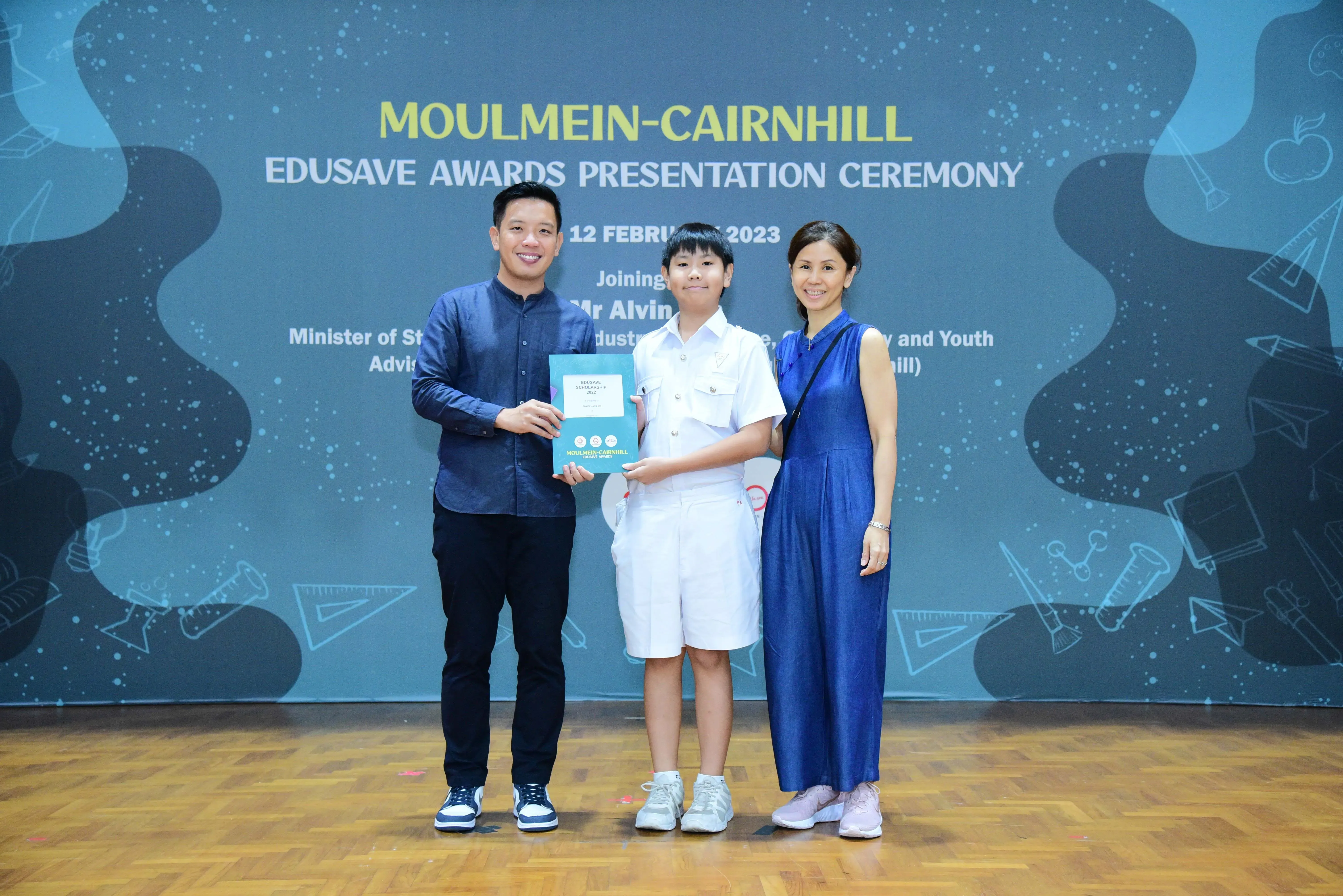 Two adults and a child in school uniform hold a certificate at the Moulmein-Cairnhill Edusave Awards Ceremony 2023.