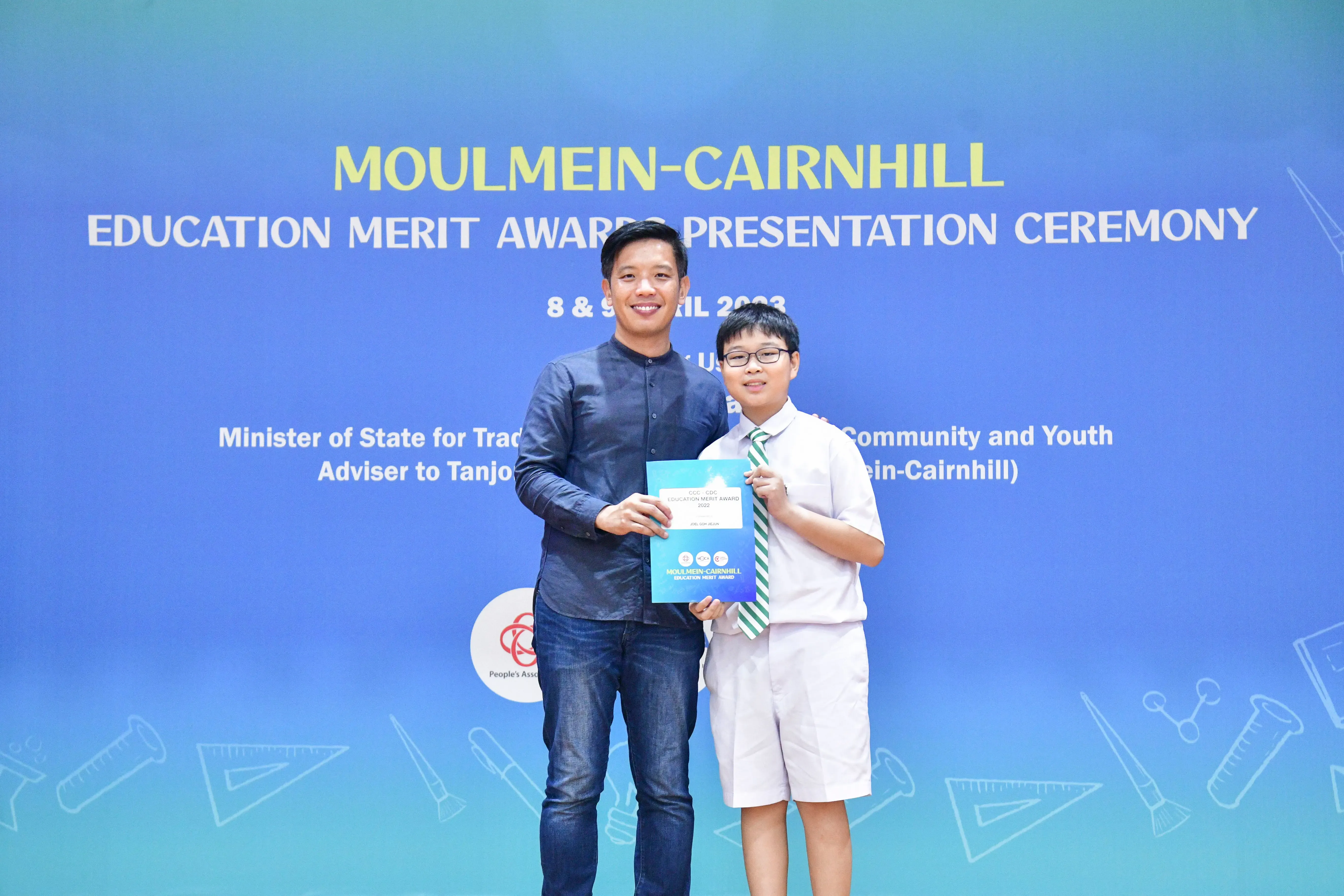 An adult and a young boy in school uniform hold a certificate at the Moulmein-Cairnhill Education Merit Awards ceremony.