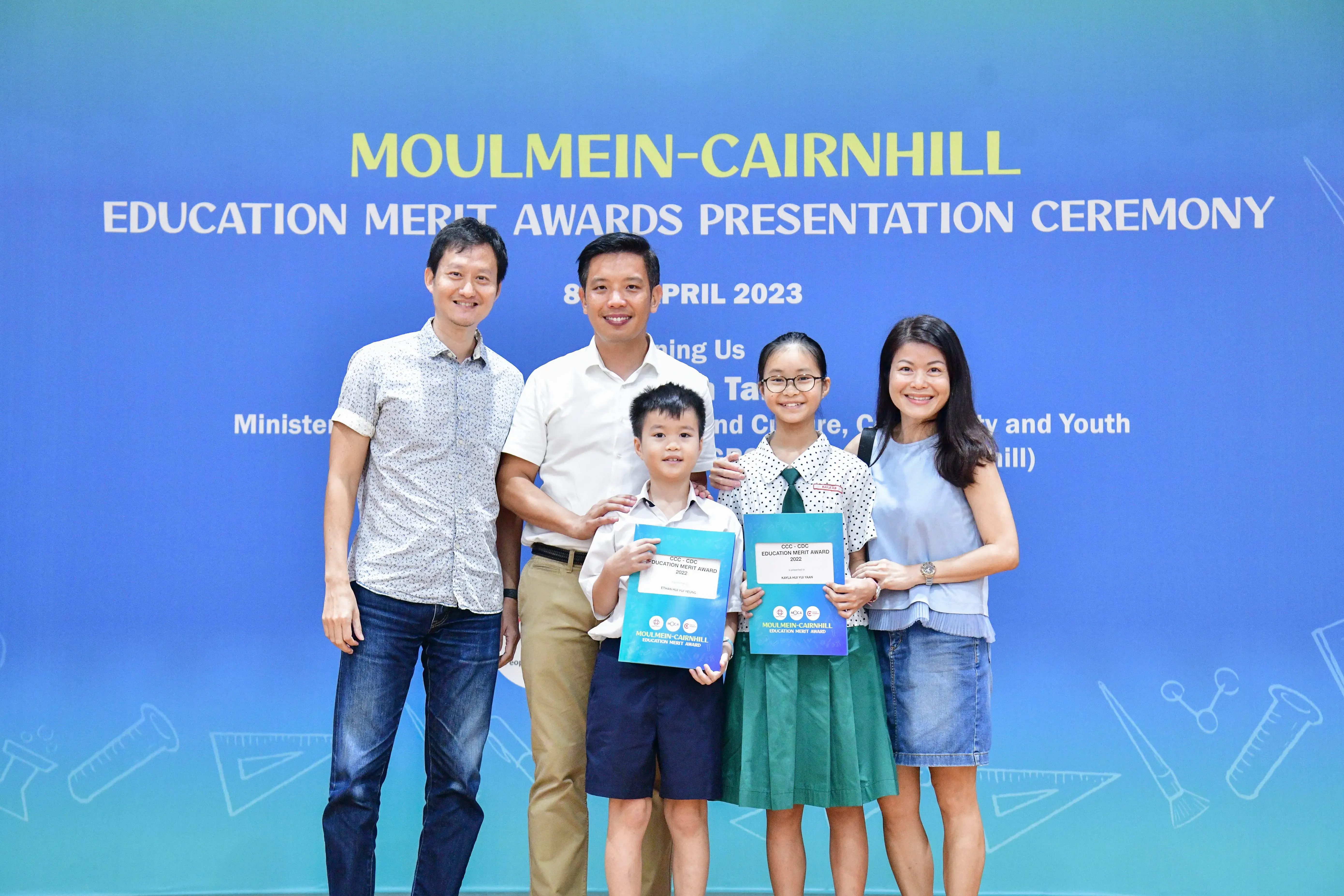 Five people pose in front of a blue Moulmein-Cairnhill Education Merit Awards Presentation Ceremony banner dated April 2023.