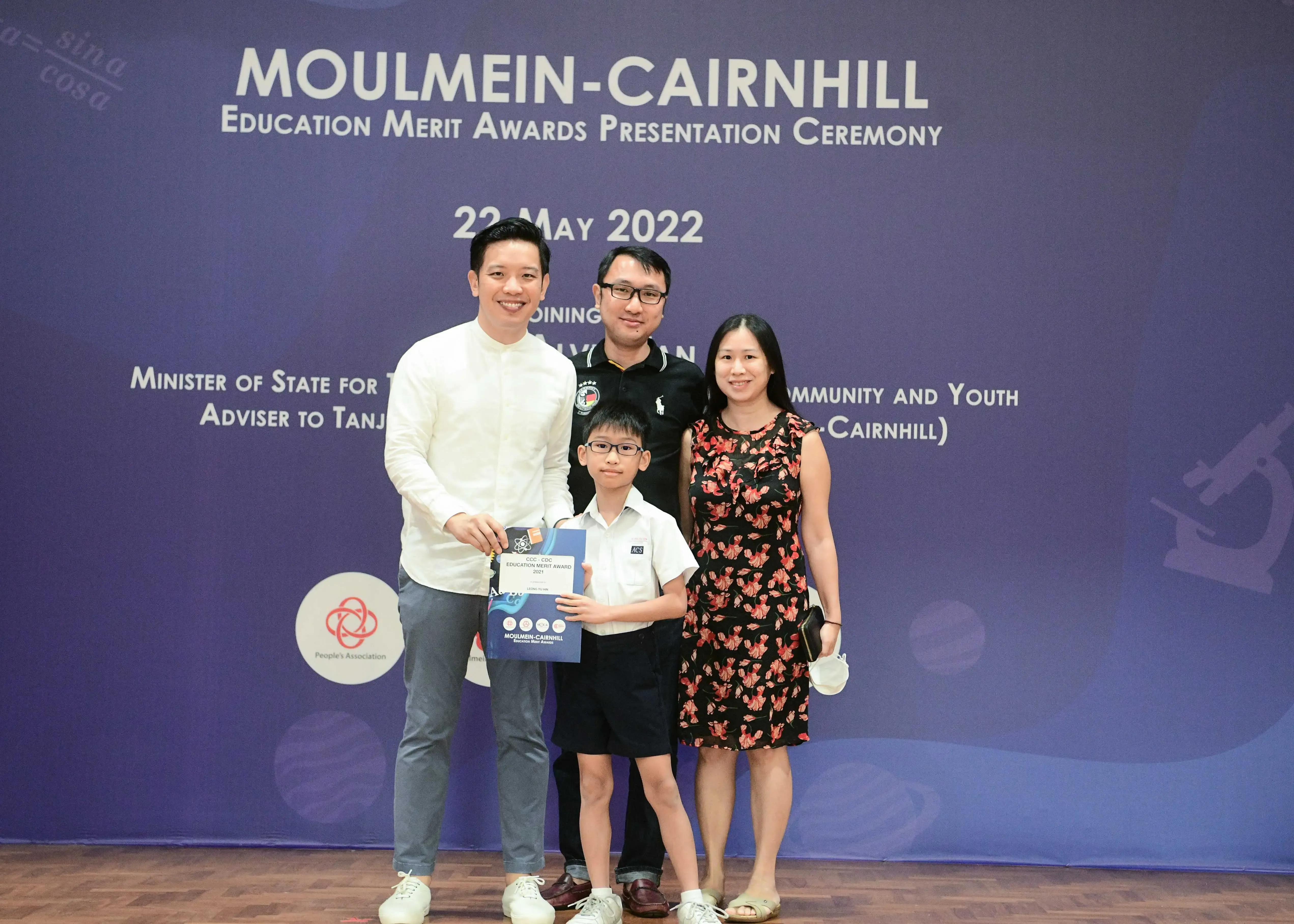 Four people pose before a Moulmein-Cairnhill Education Merit Awards banner dated 22 May 2022, with a boy holding an award.