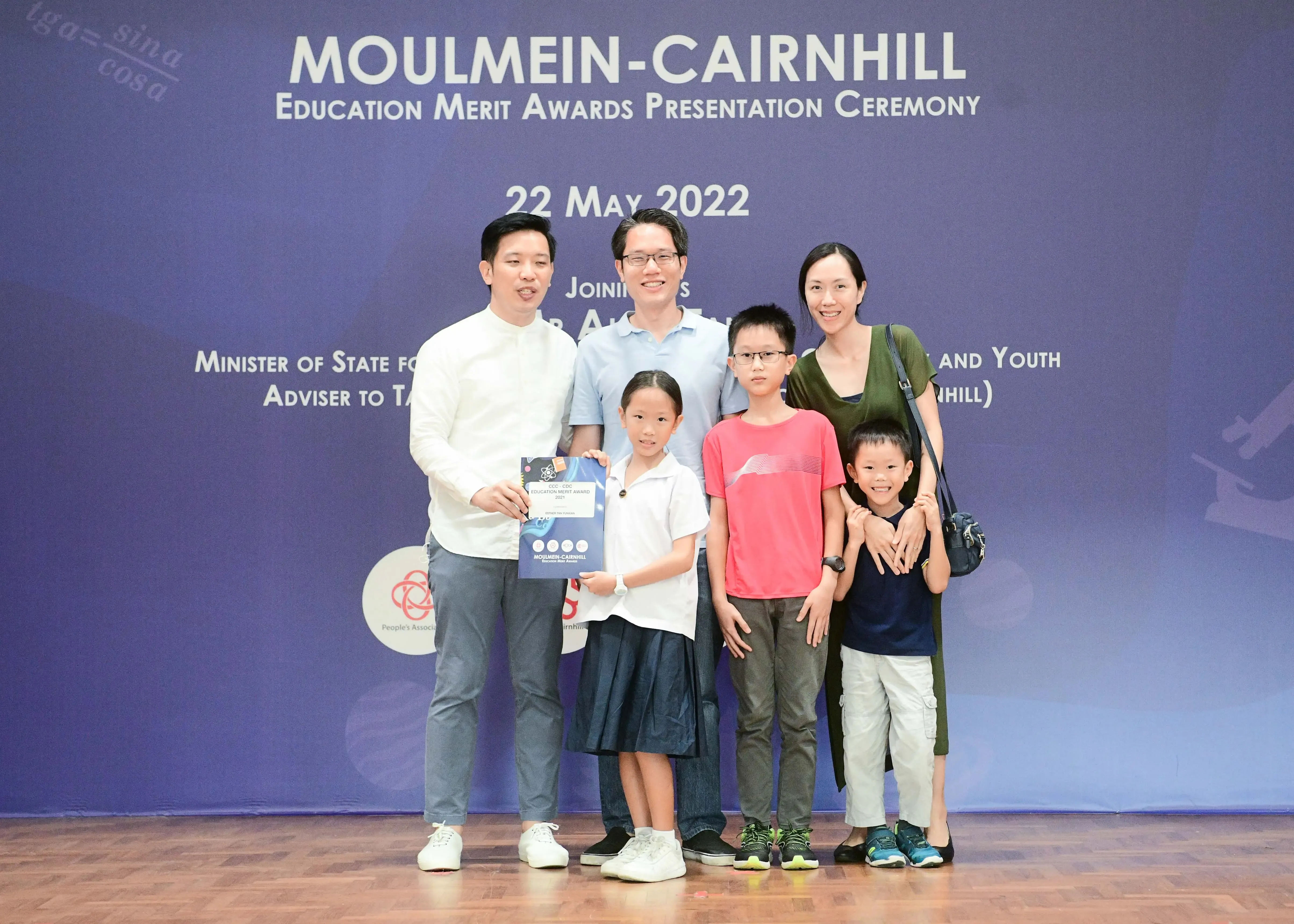 Six people pose in front of a Moulmein-Cairnhill Education Merit Awards Presentation Ceremony banner dated 22 May 2022.