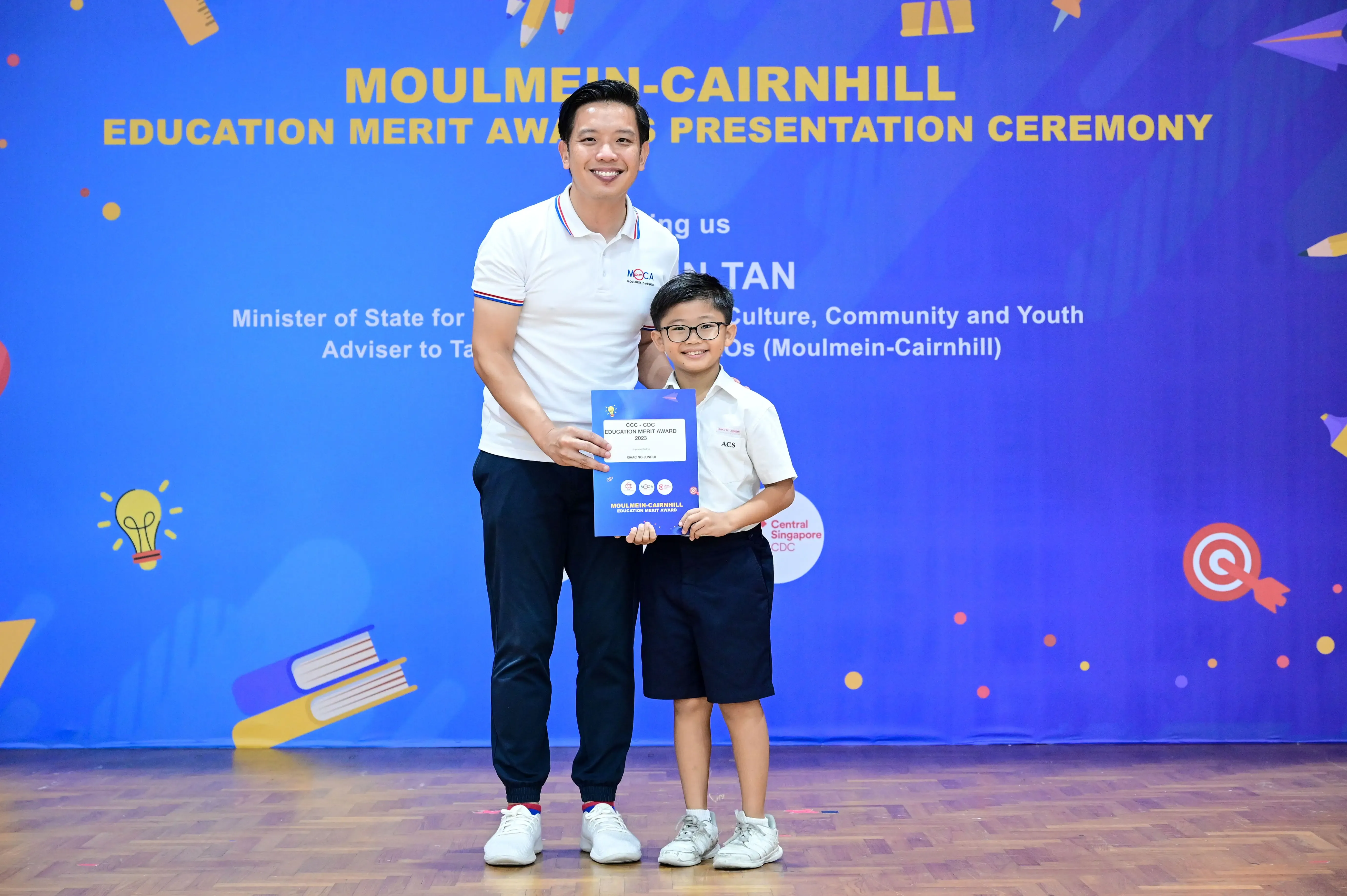 A man and a young boy in school uniform hold an award certificate at the Moulmein-Cairnhill Education Merit Award ceremony.