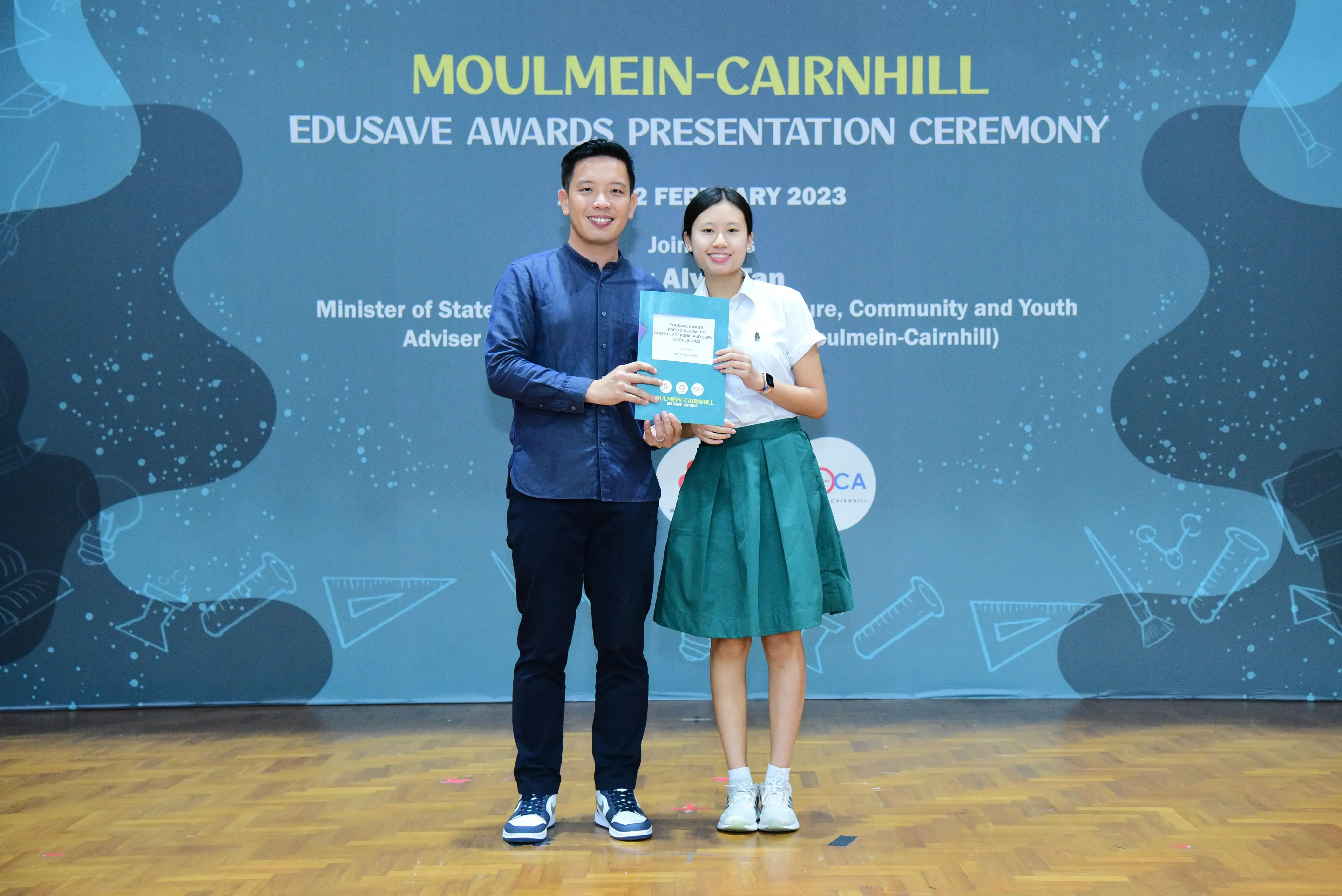Two people smile while holding a certificate at the Moulmein-Cairnhill Edusave Awards Presentation Ceremony 2023.